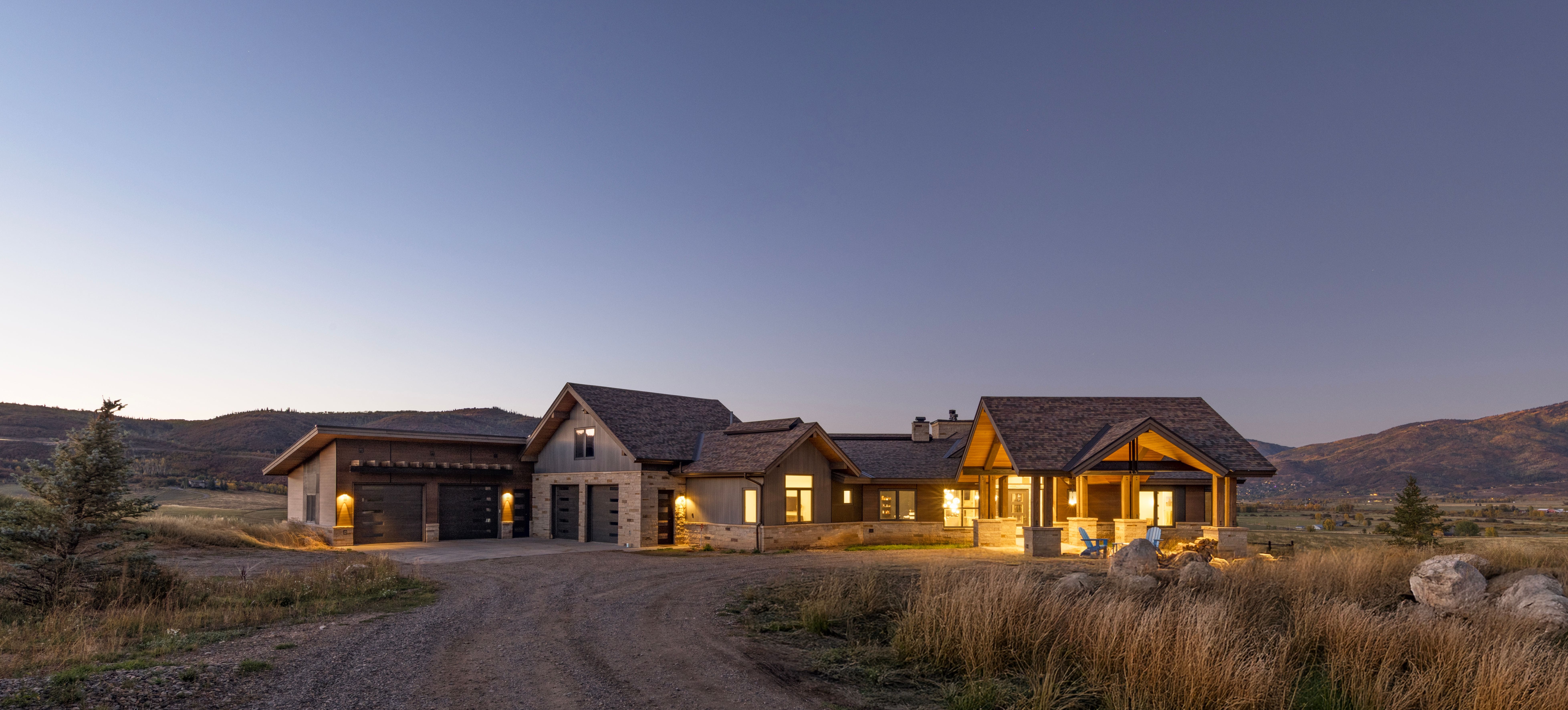 This stunning contemporary mountain estate features a harmonious blend of stone and wood siding, showcasing a multi-gabled roofline and an extensive garage complex. The exterior is beautifully illuminated, highlighting the welcoming covered entryway and a spacious covered patio overlooking the expansive, rustic landscape at twilight. The architectural design perfectly complements the serene, natural setting, offering a sophisticated and tranquil retreat.