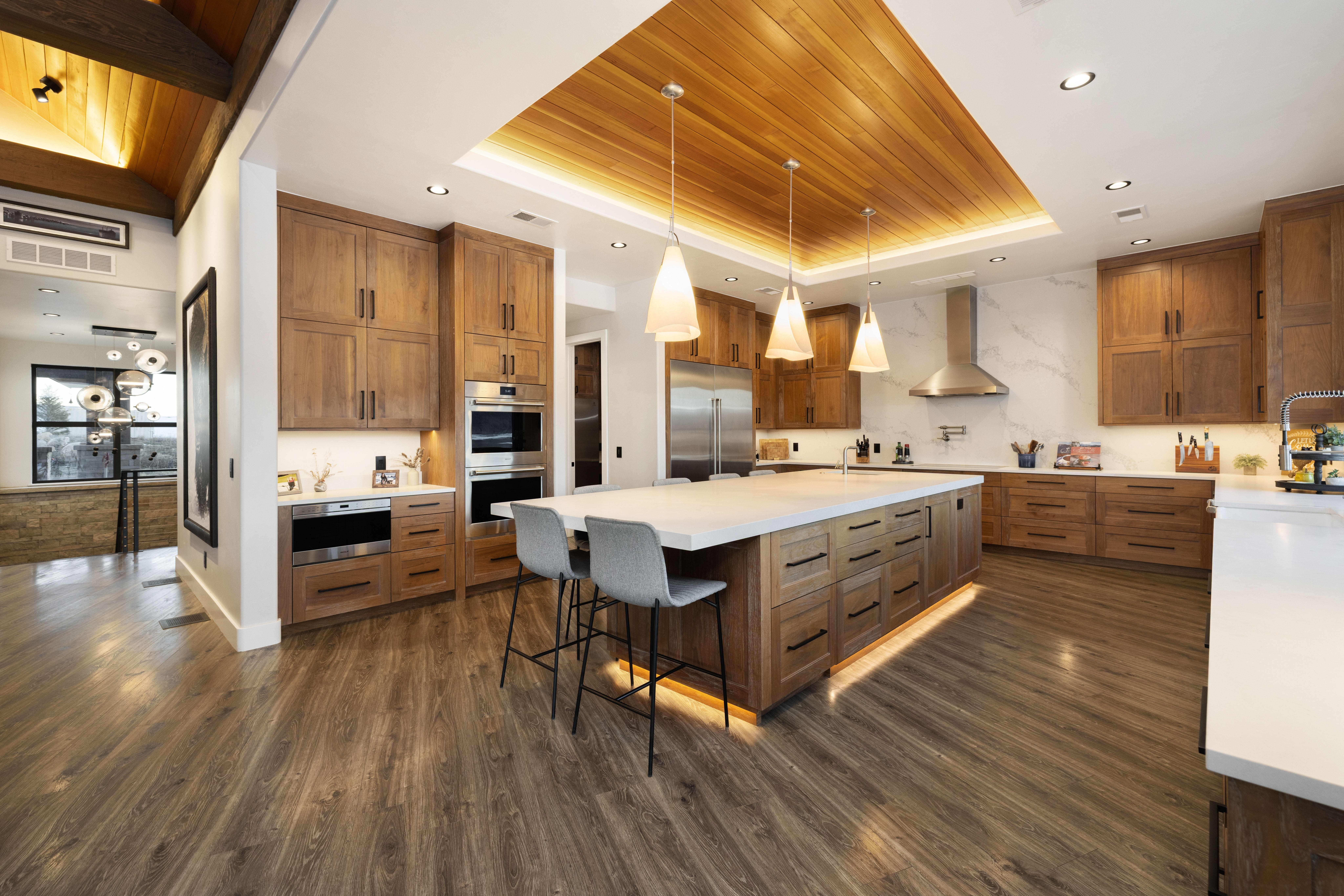 This spacious, modern kitchen features rich wood cabinetry paired with elegant white countertops and a high-end stainless steel appliance package. A large central island provides ample seating and workspace, complemented by a distinctive wood-paneled ceiling feature with integrated lighting. The open-concept design, combined with refined finishes and under-cabinet illumination, creates a warm and inviting atmosphere ideal for both cooking and entertaining.