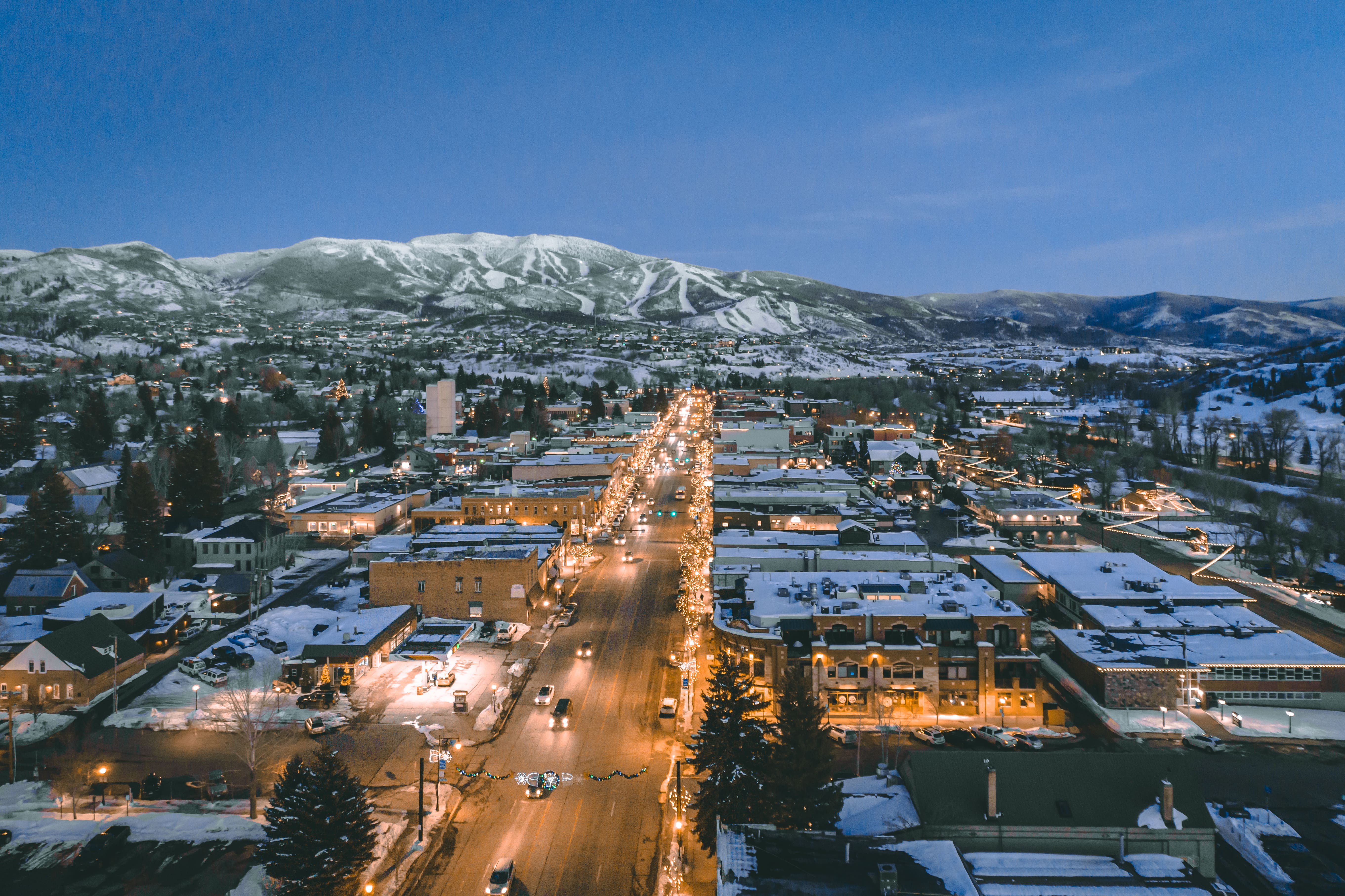 This stunning aerial view captures a charming mountain town at twilight, featuring a bustling main street illuminated by festive holiday lights. The snow-dusted landscape is set against a backdrop of majestic, expansive mountains under a serene blue sky. It provides a unique perspective on the town's central location, highlighting its picturesque atmosphere and proximity to ski terrain.
