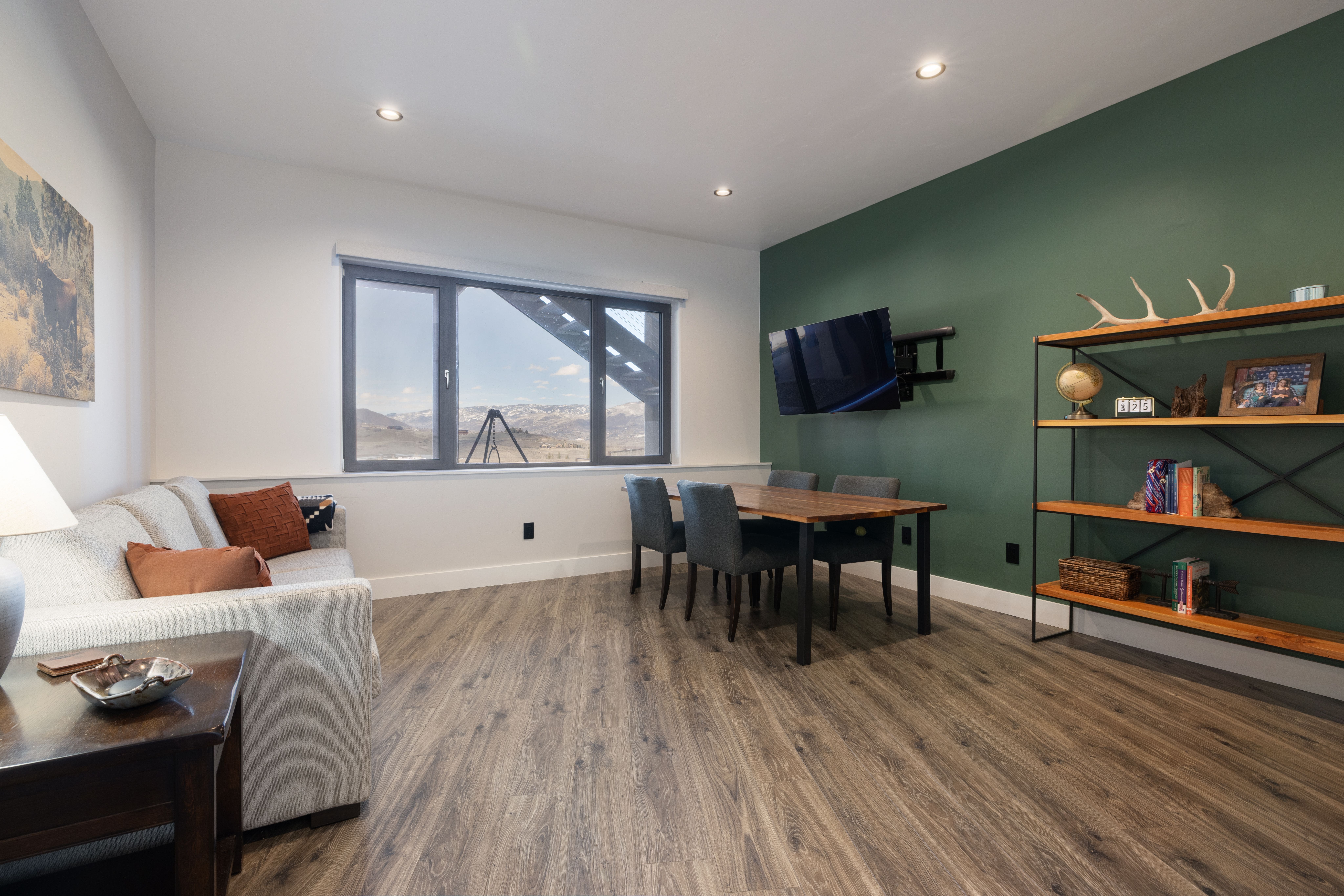 This living area features a light gray sofa with decorative accent pillows, paired with an adjacent dining space featuring a wooden table and dark gray chairs. The room is accented by a vibrant green feature wall, an open shelving unit with rustic decor, and large windows that frame expansive mountain views. Its blend of modern furniture and organic touches creates a comfortable, multi-functional space perfect for relaxing or entertaining.