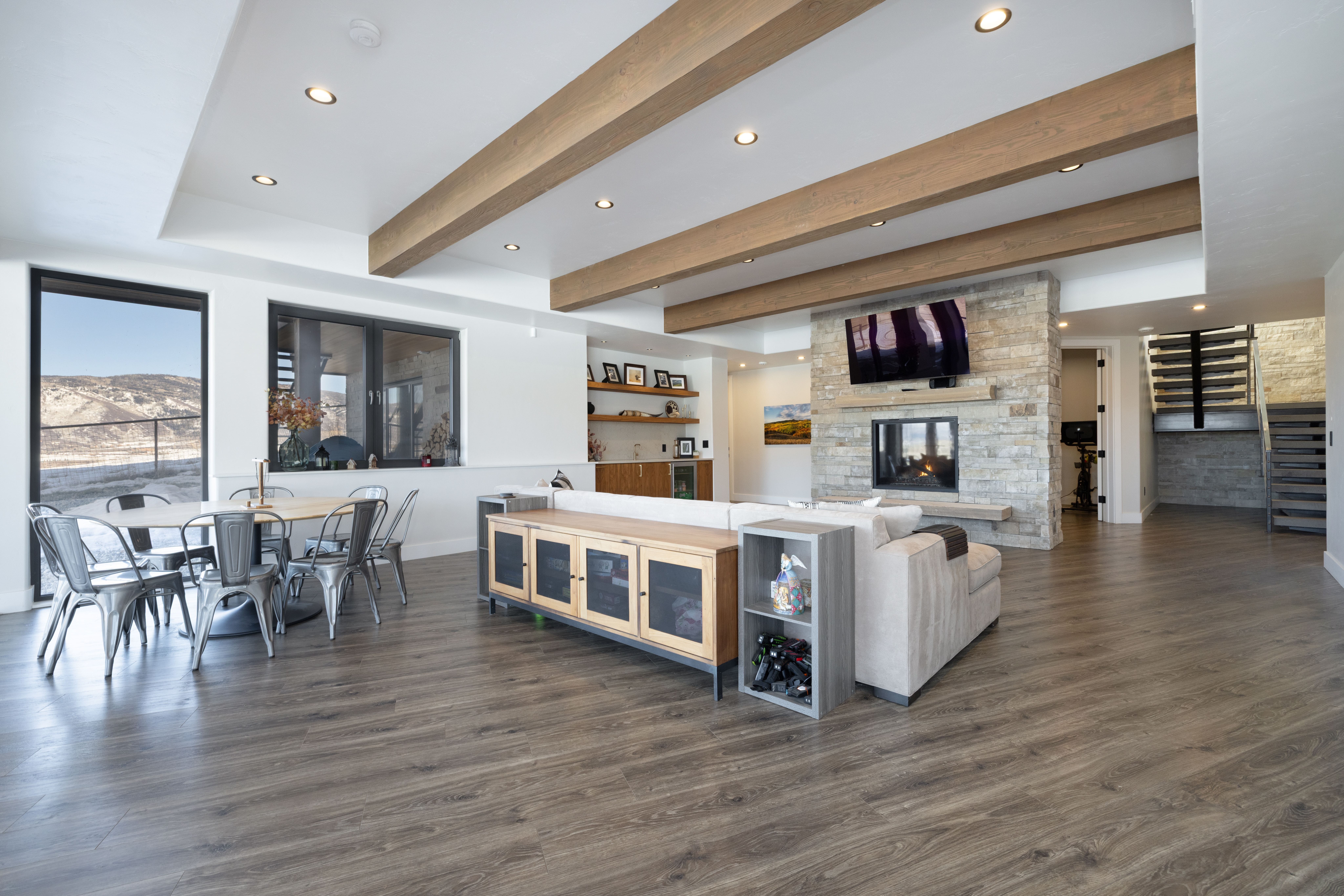 This open-concept living space features warm wood-toned flooring and a striking ceiling design with dark-stained exposed timber beams. The focal point is a floor-to-ceiling stone fireplace with a built-in television, complemented by a cozy sectional sofa and a custom wooden console table. An adjacent dining area with metal chairs and large windows creates a seamless, modern aesthetic with plenty of natural light.