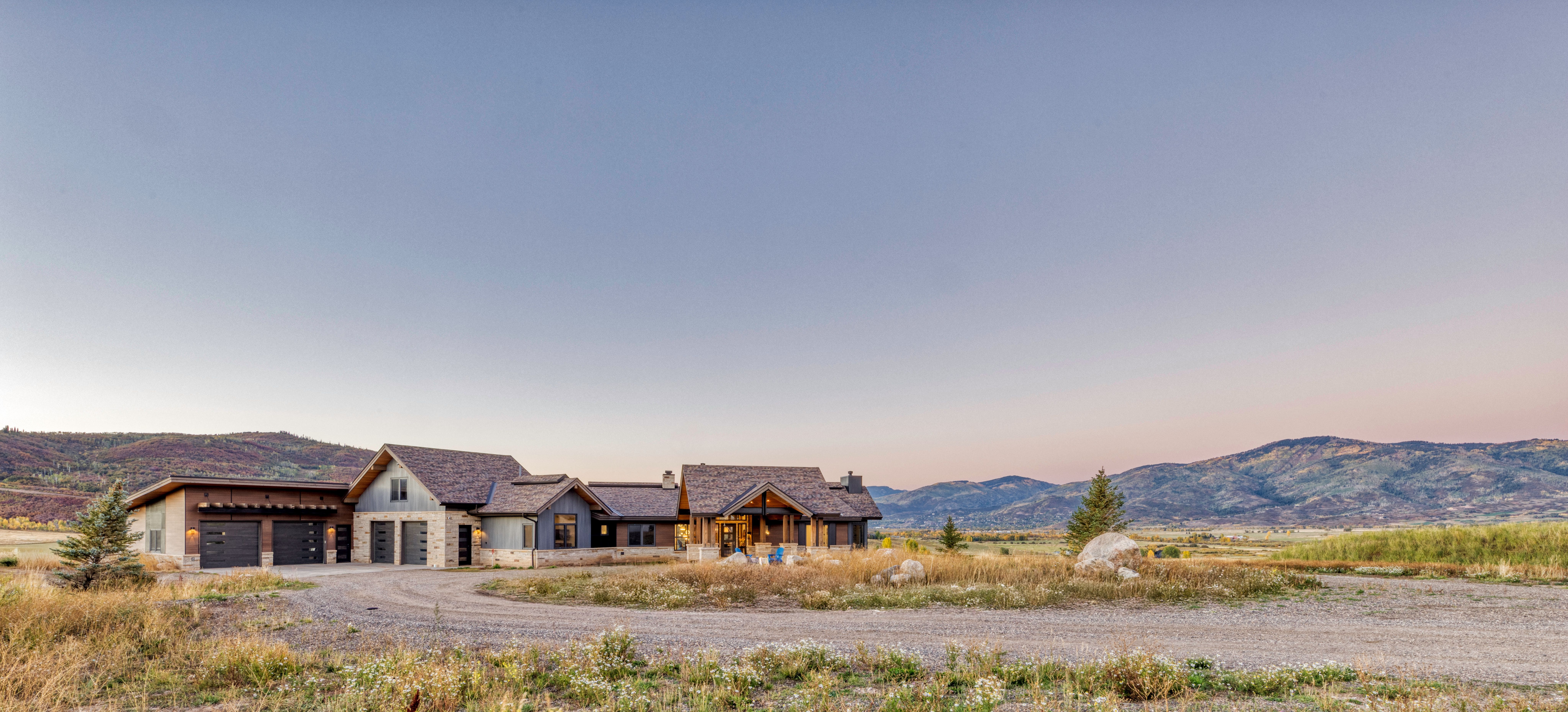 This stunning contemporary mountain estate features a harmonious blend of stone and wood siding, complemented by a multi-gabled roofline. Set against a backdrop of sweeping mountain vistas and vast, natural wild grass fields, the home offers a serene and expansive rural living experience. The wide gravel driveway leads to a multi-car garage, anchoring a residence that seamlessly integrates luxury architectural design with the rugged beauty of the surrounding landscape.