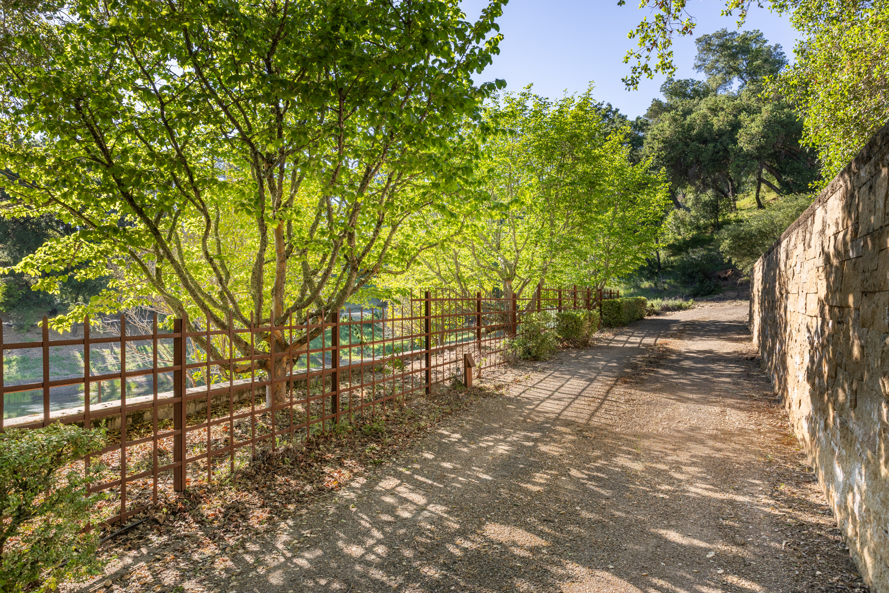 This serene outdoor pathway features a rustic gravel trail bordered by a tall, textured stone wall on one side and a decorative metal trellis fence on the other. Lush, mature trees provide dappled shade across the path, creating a peaceful and inviting atmosphere. The perspective captures the depth of the garden, highlighting the blend of natural landscaping and structured architectural elements.