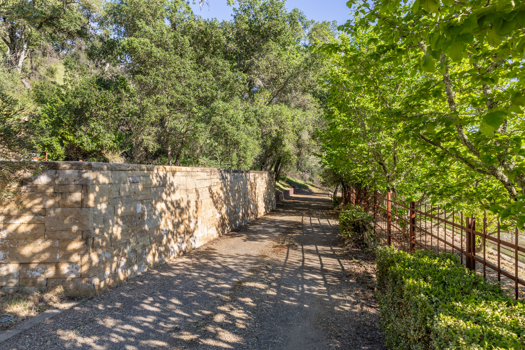 This image captures a serene, sun-dappled pathway lined by a rustic stone retaining wall on the left and a metal fence bordered by lush greenery on the right. The perspective looks down the gravel path, which is framed by mature trees that cast intricate shadows across the ground, creating a peaceful and secluded atmosphere. The scene highlights the property's natural landscaping and well-maintained outdoor features, suggesting a quiet, private retreat.