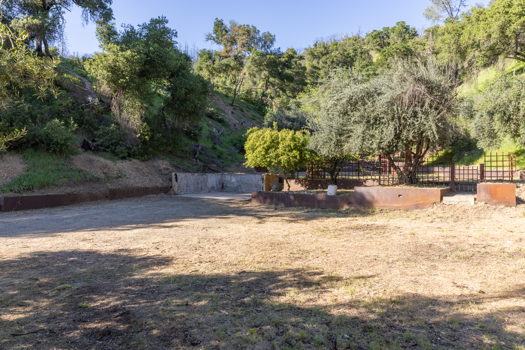 This outdoor space features a large, dry dirt lot bordered by rustic, weathered steel retaining walls that create a tiered landscape effect. The area is framed by mature trees and lush hillside vegetation, offering a natural, secluded setting with a sense of rustic charm. The perspective is a wide, eye-level shot that captures the expansive potential of the grounds and the integration of the man-made structures with the surrounding terrain.