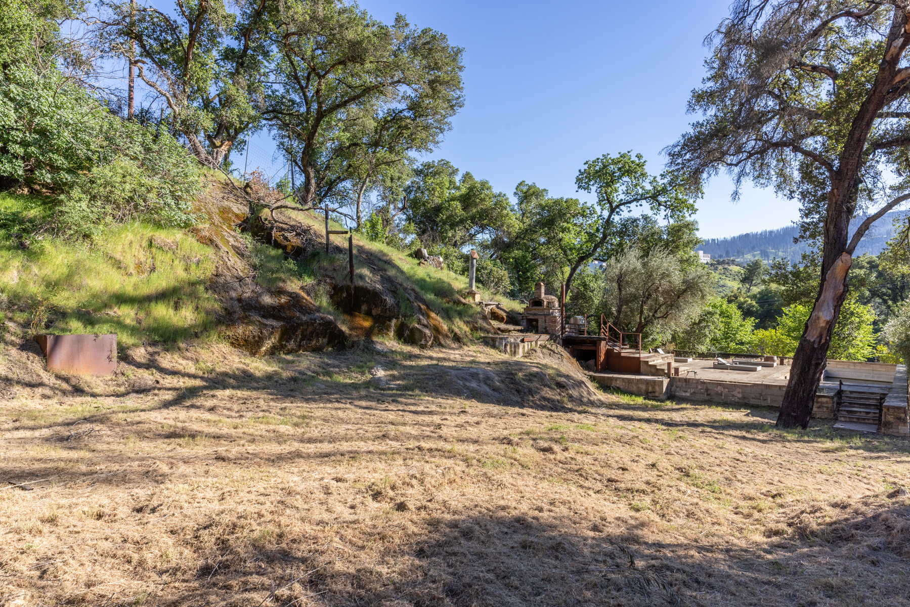 This expansive, sun-drenched outdoor space features a large, dry grassy slope leading up to a rugged, rocky hillside dotted with mature oak trees. A rustic brick outdoor oven and a tiered stone patio area with a pool are nestled into the landscape, offering a serene and natural setting. The perspective captures the vastness of the property, highlighting its potential for outdoor entertaining and peaceful rural living.