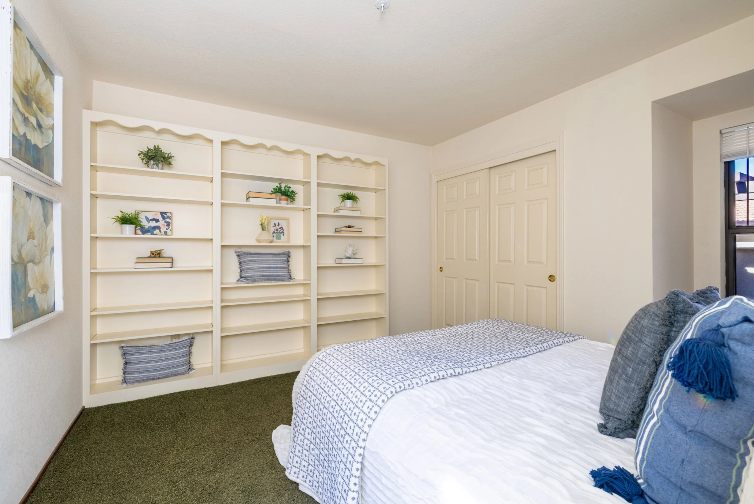 This cozy bedroom features a neutral color palette with a prominent built-in shelving unit that adds character and storage space. The room is furnished with a comfortable bed dressed in a patterned blue and white throw, complemented by matching decorative pillows. The perspective is from the foot of the bed, looking toward the built-in shelves and the closet, creating a warm and inviting atmosphere.