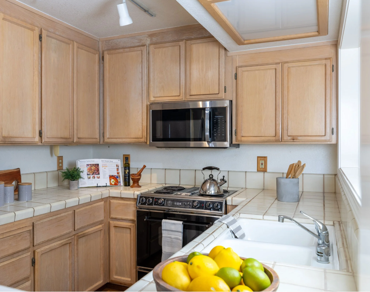 This kitchen features light oak cabinetry, tiled countertops, and a stainless steel microwave and stove, offering a classic, functional aesthetic. A bowl of fresh lemons and limes sits in the foreground near the white double-basin sink, while a cookbook and mortar and pestle add a lived-in, inviting touch. The perspective is a warm, eye-level shot that highlights the efficient layout and cozy atmosphere of the space.