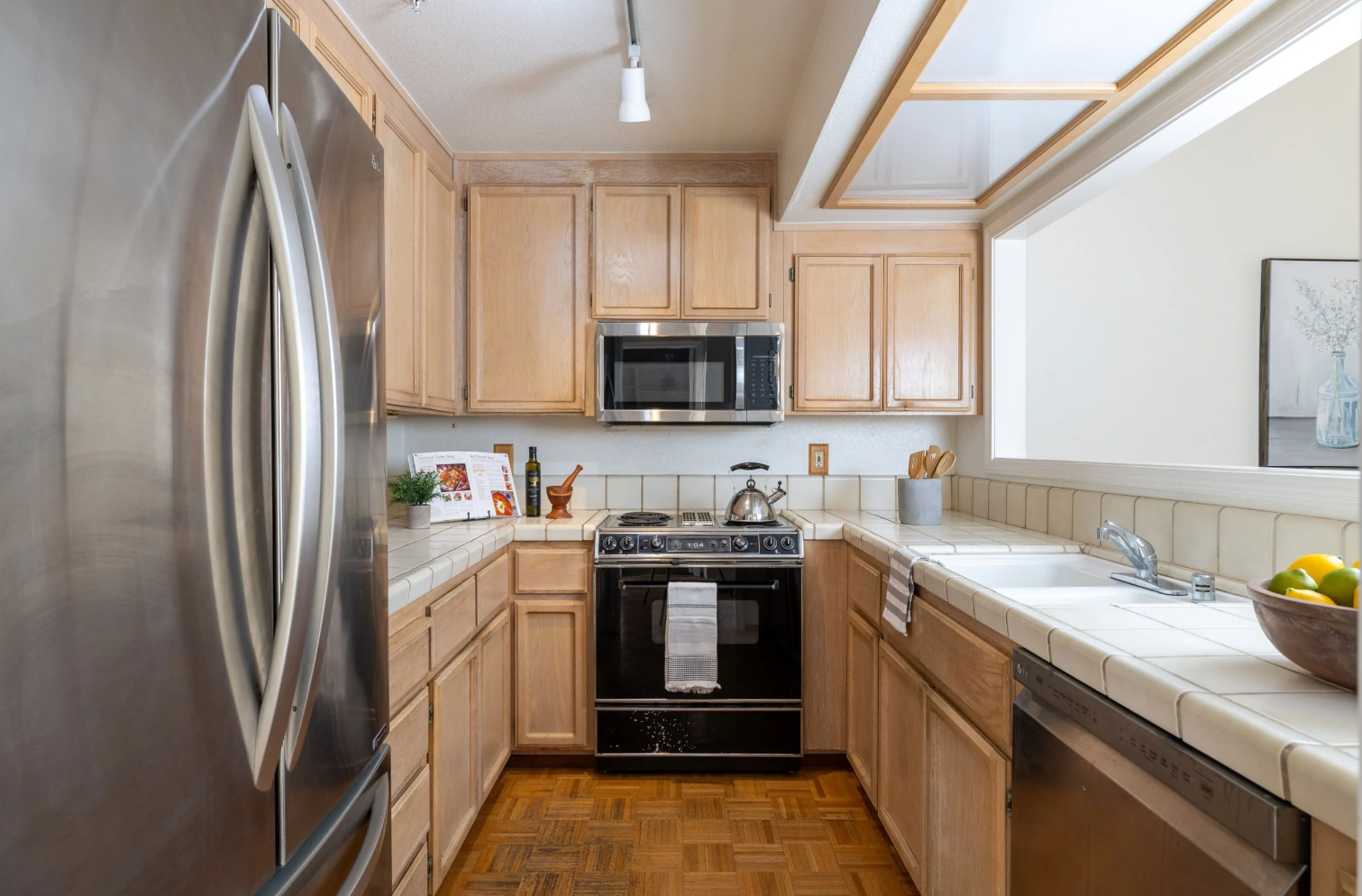 This U-shaped kitchen features light oak cabinetry, a stainless steel refrigerator, and a black range with a matching microwave above. The space is finished with tiled countertops and a classic parquet wood floor, creating a warm and functional atmosphere. The perspective is a straight-on, eye-level shot that highlights the efficient layout and the pass-through window looking into the adjacent room.