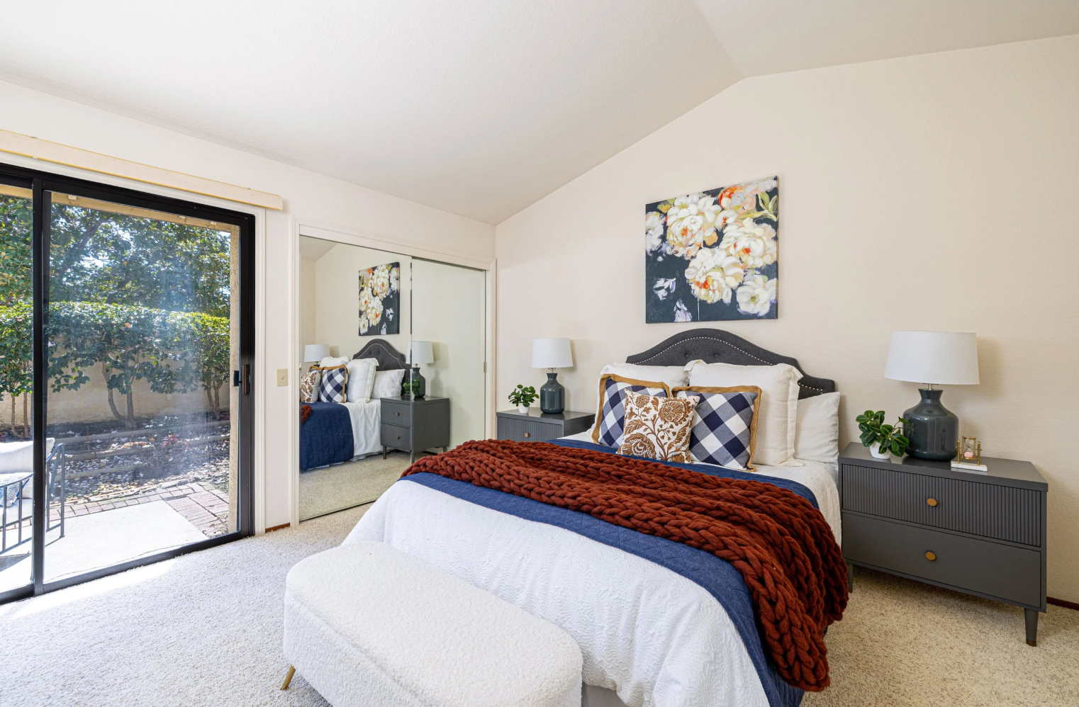 This cozy primary bedroom features a queen-sized bed adorned with a chunky rust-colored knit throw and a mix of patterned pillows, set against a neutral wall with floral artwork. A large sliding glass door provides ample natural light and a view of the outdoor patio, while mirrored closet doors enhance the sense of space. The room is completed by matching dark grey nightstands and a plush white bench at the foot of the bed, creating a warm and inviting atmosphere.
