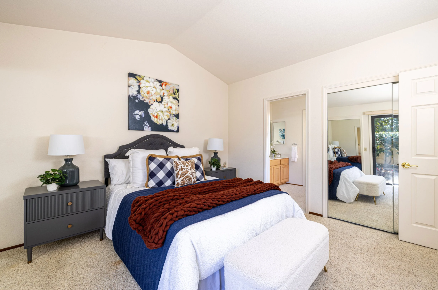 This cozy primary bedroom features a dark grey upholstered headboard paired with a navy blue quilt and a chunky, rust-colored knit throw blanket. The room is accented by matching dark grey nightstands, a white textured bench at the foot of the bed, and a floral art piece above the headboard. A large mirrored closet door reflects the space, adding depth and brightness to the room, which also includes a view into an adjoining bathroom.