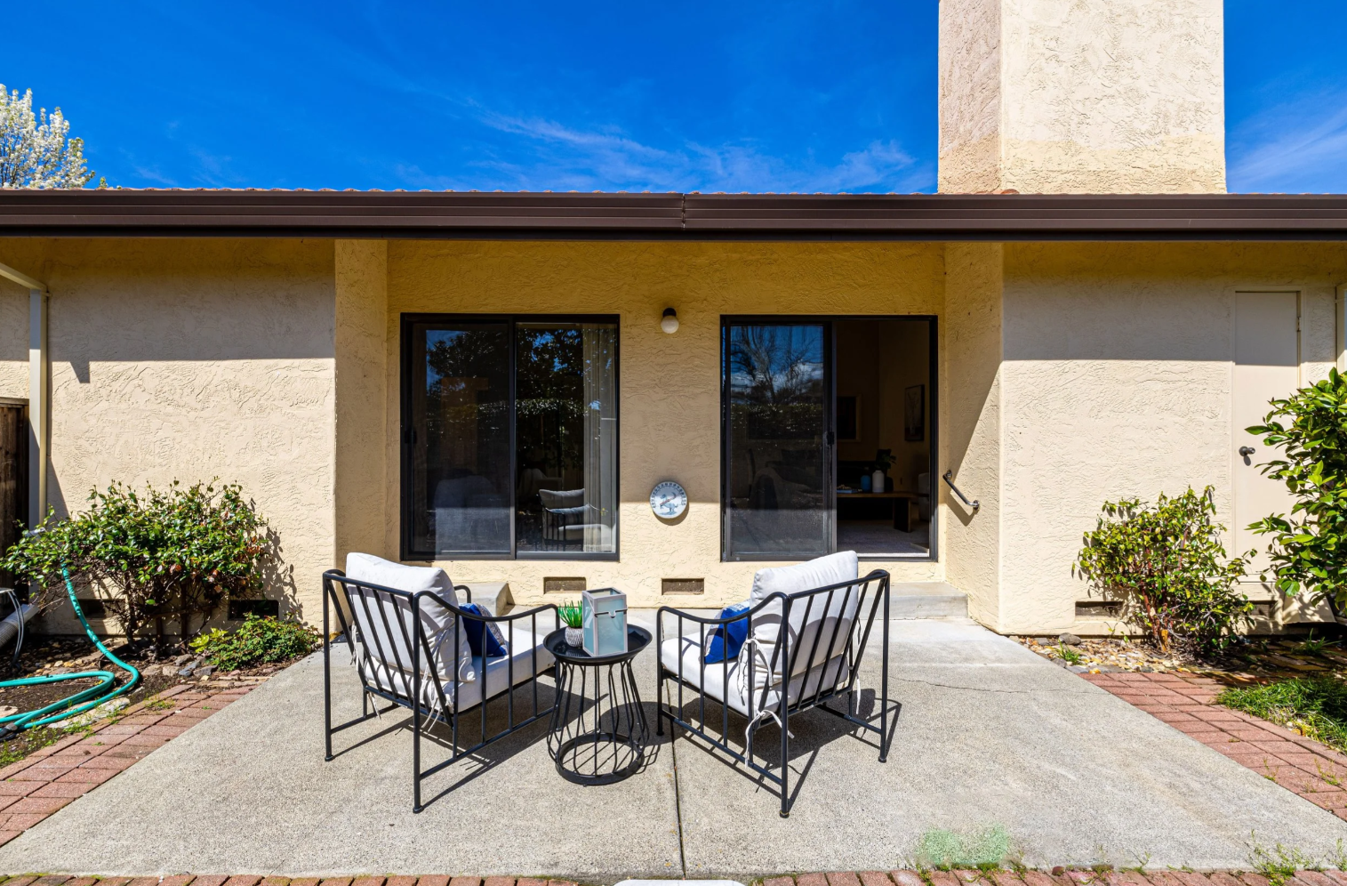 This image showcases a clean, concrete patio area featuring two modern black-framed chairs with white cushions and a small matching side table. The patio is situated directly against the rear of a light-yellow stucco home, providing access through two sliding glass doors. The scene is bathed in bright, natural sunlight under a clear blue sky, creating an inviting and peaceful outdoor living space.