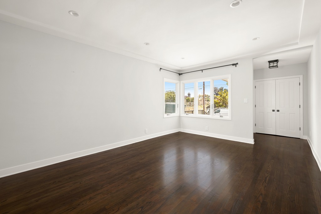 This bright, unfurnished room features dark hardwood flooring and neutral light-gray walls, creating a clean and versatile living space. A corner window arrangement allows for natural light to fill the room, while a set of white double doors provides access to a closet or adjacent area. The perspective is wide-angle, capturing the entire room's layout and emphasizing its spacious, minimalist aesthetic.