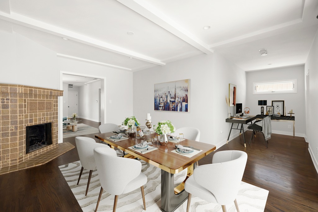 This open-concept dining area features a modern wooden table surrounded by six white upholstered chairs, set atop a patterned area rug. To the left, a fireplace with a textured tile surround adds a focal point, while the right side of the room transitions into a dedicated home office space with a sleek desk. The room is bright and airy, with dark wood flooring and a neutral color palette that creates a sophisticated and inviting atmosphere.