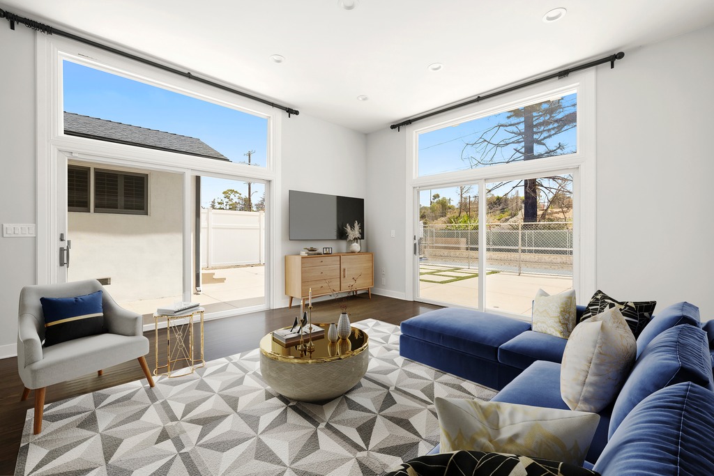 This bright and airy living room features a contemporary design with a large blue sectional sofa and a matching armchair. The space is anchored by a geometric patterned area rug and a circular gold-rimmed coffee table, while large sliding glass doors provide ample natural light and views of the outdoor patio. A television is mounted above a light wood media console, creating a clean and inviting focal point for the room.