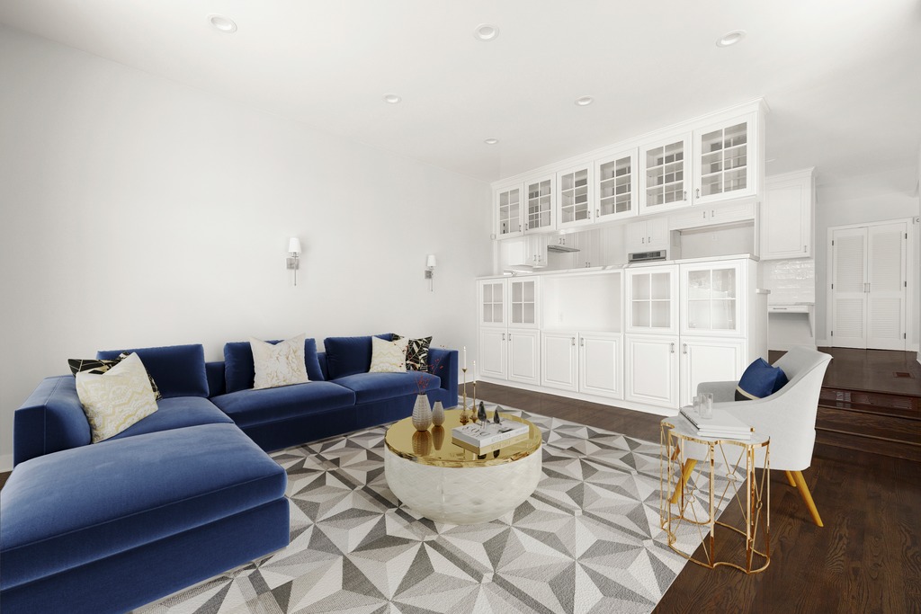 This bright and airy living room features a large, plush blue sectional sofa accented with patterned throw pillows, centered around a round coffee table with a gold base and white top. The space is anchored by a geometric-patterned area rug and includes a white armchair with a matching gold-accented side table. In the background, a wall of white cabinetry with glass-front doors provides ample storage and display space, creating a clean, sophisticated aesthetic.
