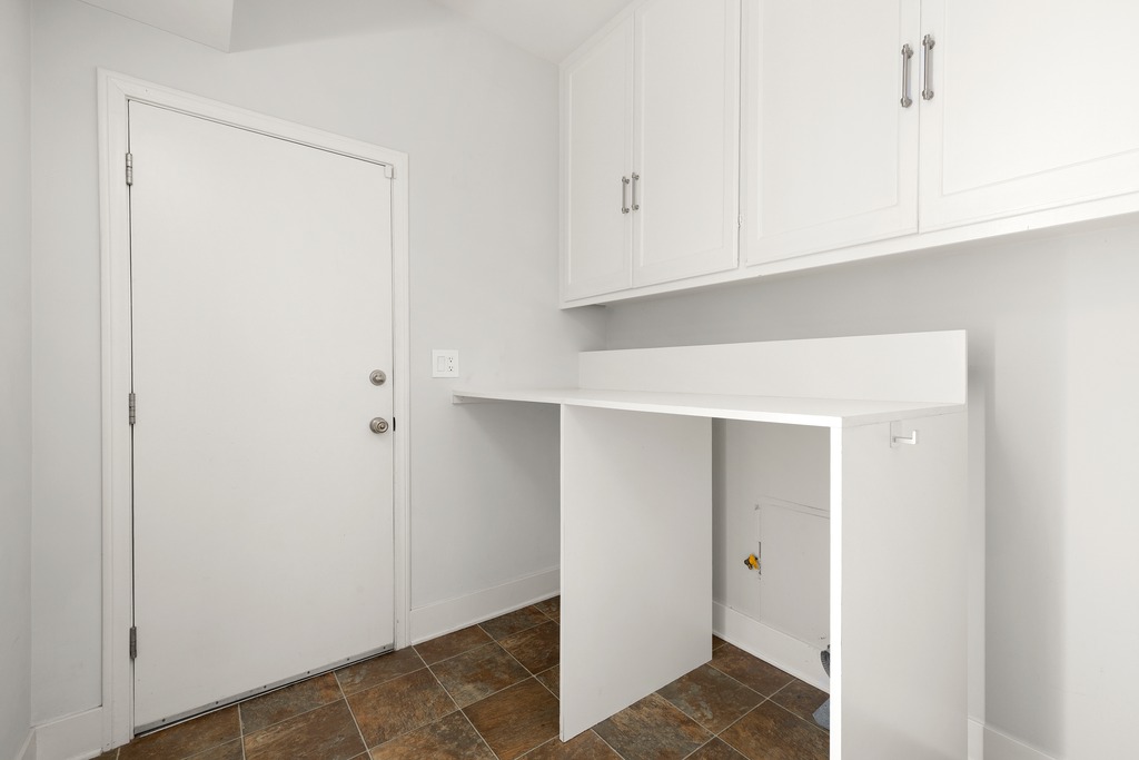 This laundry room features a clean, minimalist aesthetic with white cabinetry and a matching white countertop workspace. The room includes a door leading to the exterior and patterned tile flooring, providing a functional and bright utility space. The perspective is a straight-on view, highlighting the ample storage and dedicated area for laundry appliances.