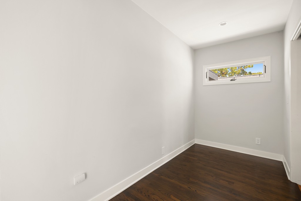 This minimalist room features dark hardwood flooring and light gray walls, creating a clean and neutral backdrop. A single horizontal window is positioned high on the wall, allowing natural light to enter while maintaining privacy. The perspective is from the corner of the room, highlighting the simple, modern aesthetic of the space.