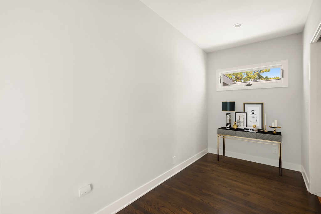 This minimalist space features dark hardwood flooring and neutral light-colored walls, creating a clean and modern aesthetic. A sleek console table with gold-toned legs serves as a focal point, adorned with decorative items and a small lamp beneath a horizontal window. The perspective is a wide-angle shot from the corner of the room, highlighting the open floor space and the simple, elegant decor.