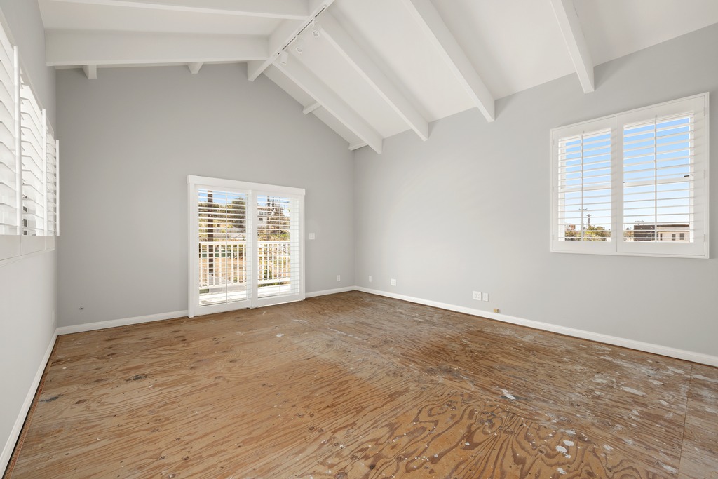 This spacious, unfurnished room features a high vaulted ceiling with exposed white beams and light gray walls. The space is illuminated by natural light streaming through a large window with white plantation shutters and a set of sliding glass doors leading to an outdoor area. The floor is currently unfinished plywood, presenting a blank canvas ready for new flooring installation.