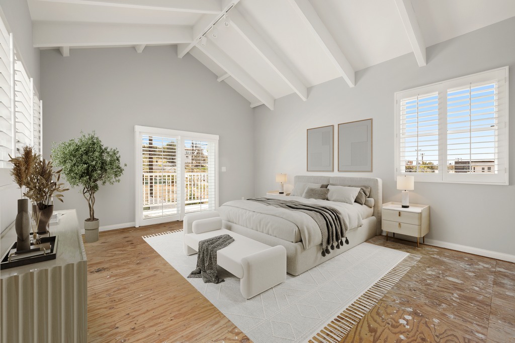 This spacious primary bedroom features a vaulted ceiling with exposed white beams and light-toned wood flooring. The room is anchored by a neutral-colored bed with a matching bench at the foot, flanked by two modern nightstands with lamps. Large windows and a glass door with plantation shutters allow for ample natural light, creating an airy and serene atmosphere.
