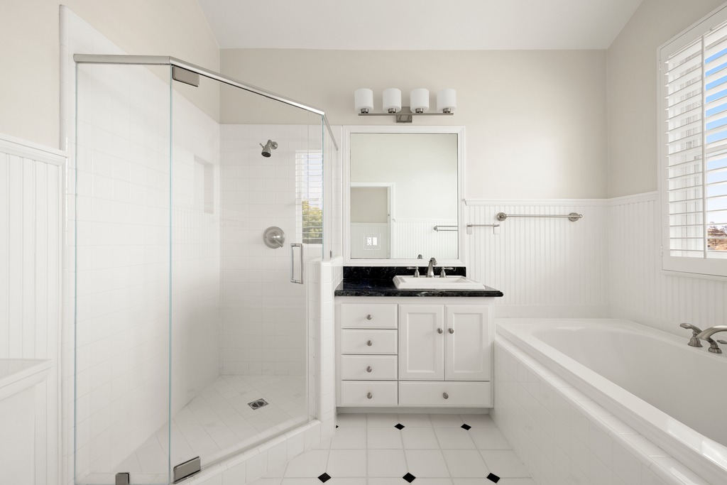 This bright and clean bathroom features a corner glass-enclosed shower, a white vanity with a dark stone countertop, and a deep soaking tub. The space is finished with white beadboard wainscoting and white floor tiles accented with small black diamond inlays. The perspective is a straight-on shot capturing the functional layout of the vanity, shower, and tub area.