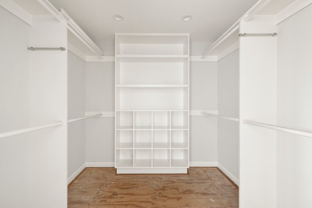 This walk-in closet features a symmetrical layout with white built-in shelving and hanging rods on both sides. A central unit provides open shelving and a grid of cubbies for organized storage. The space is currently unfinished, with exposed plywood flooring, offering a blank canvas for customization.