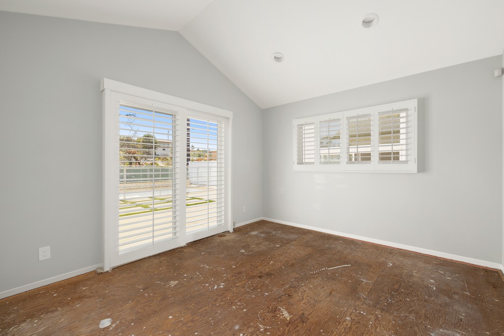 This bright, empty room features light gray walls and a vaulted ceiling, creating an airy and spacious atmosphere. The space includes a set of glass sliding doors and a large window, both fitted with white plantation shutters that allow for adjustable natural light. The floor is currently unfinished plywood, presenting a blank canvas ready for new flooring installation.