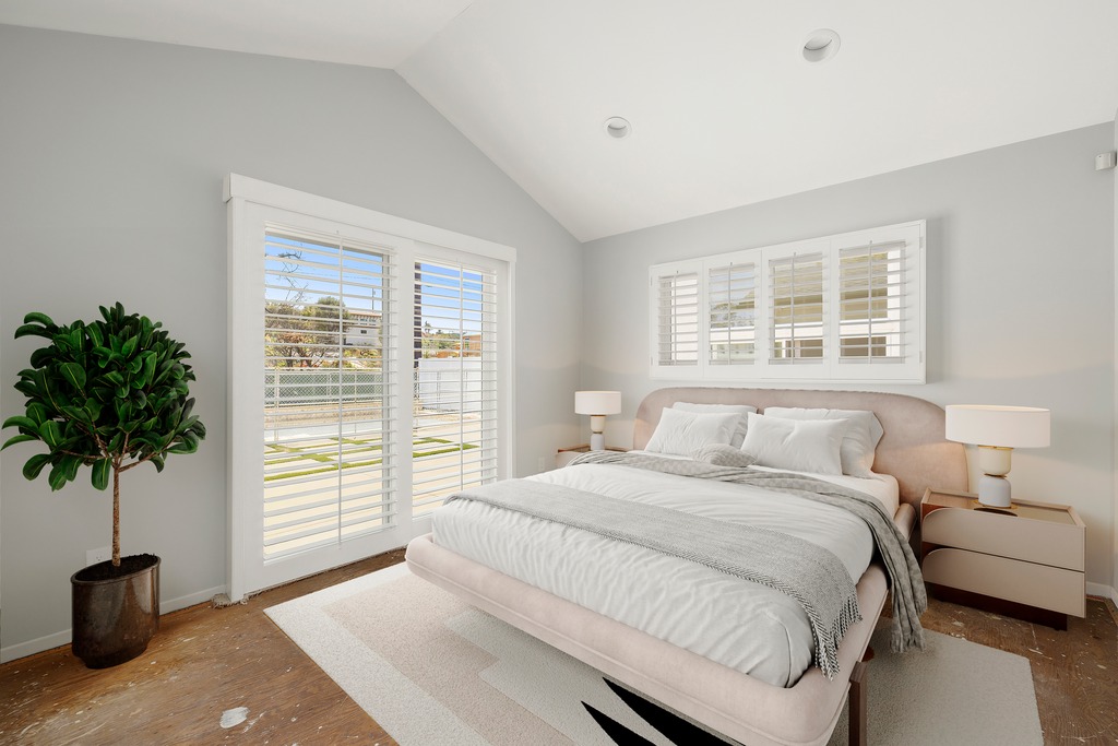 This primary bedroom features a vaulted ceiling and a neutral color palette, creating an airy and serene atmosphere. A plush, upholstered bed serves as the focal point, flanked by two modern nightstands with minimalist lamps. Large glass doors with plantation shutters allow for natural light, while a potted plant adds a touch of greenery to the space.