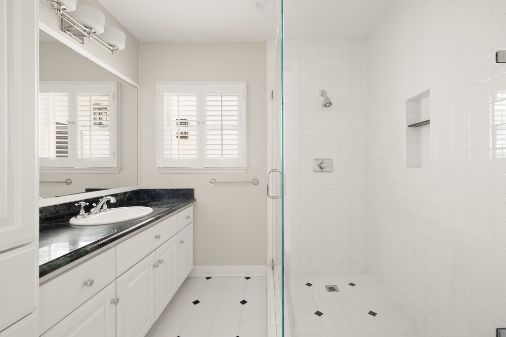 This bright and clean bathroom features a white vanity topped with a dark, polished stone counter and a classic undermount sink. A glass-enclosed shower with white wall tiles and a built-in niche sits to the right, while the floor is finished with white tiles accented by small black diamond inlays. The space is illuminated by a multi-bulb vanity light above a large mirror and natural light streaming through plantation-shuttered windows.