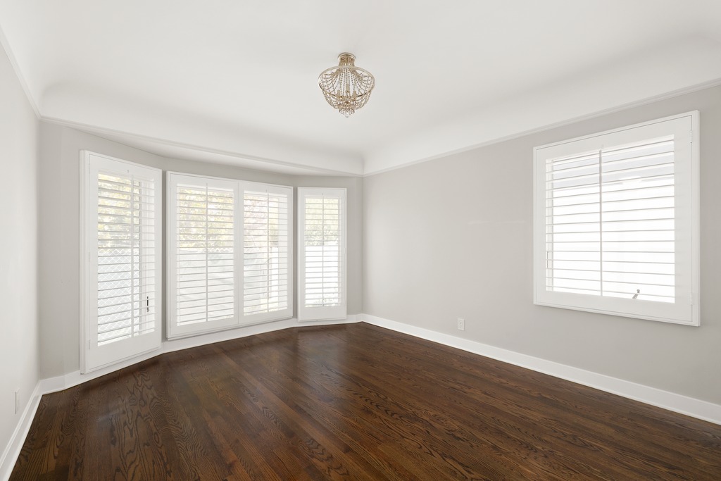 This bright and airy room features dark wood flooring and neutral light gray walls, creating a clean and versatile living space. A bay window area with white plantation shutters allows for ample natural light, complemented by a single window on the adjacent wall and a decorative crystal chandelier. The perspective is captured from a low angle, emphasizing the spaciousness and the contrast between the dark floors and light walls.