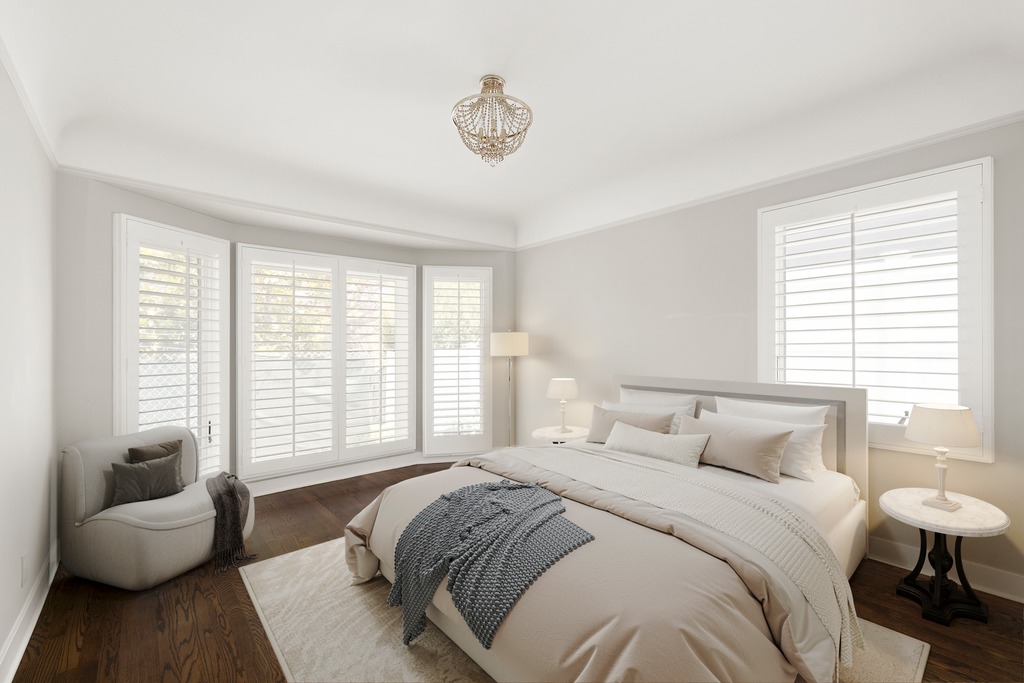This serene primary bedroom features a neutral color palette, dark hardwood flooring, and a plush bed dressed in layered linens and a textured throw. The room is illuminated by a decorative crystal chandelier and soft bedside lamps, while large windows with white plantation shutters allow for adjustable natural light. A comfortable armchair sits in the corner, creating a cozy reading nook within this elegant and well-appointed space.