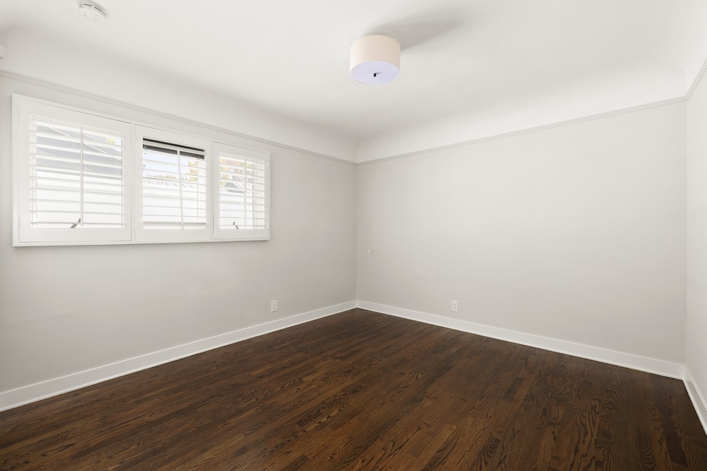 This bright, empty bedroom features dark hardwood flooring and neutral light-colored walls, creating a clean and versatile living space. A large, multi-pane window with white plantation shutters allows for natural light, while a simple drum-style ceiling fixture provides overhead illumination. The room is framed by crisp white baseboards and crown molding, offering a classic and well-maintained aesthetic.