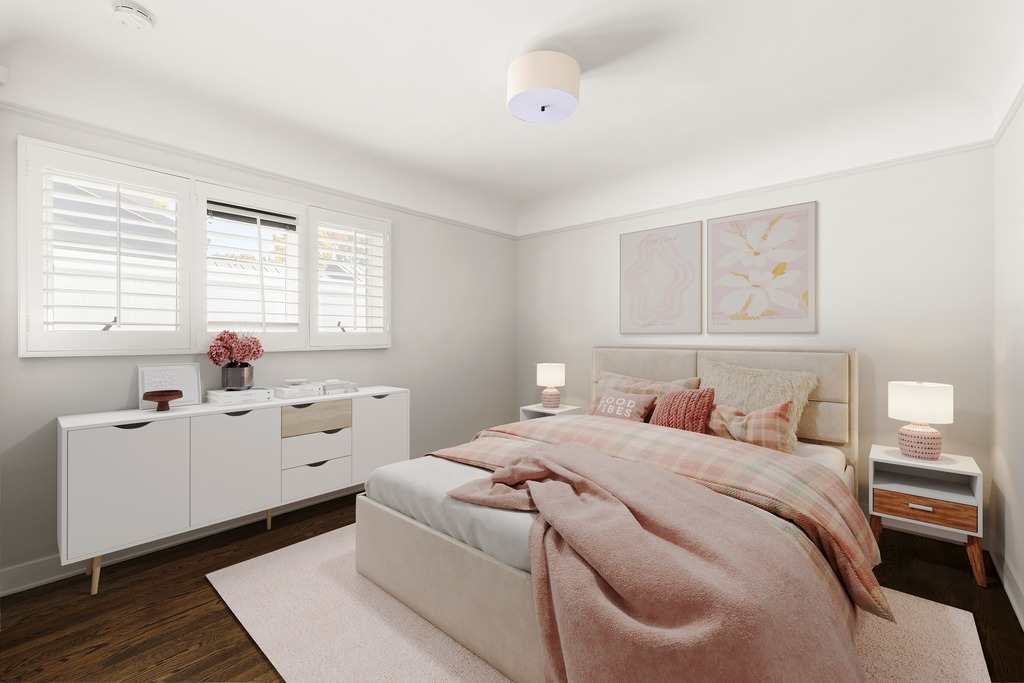 This bright and inviting bedroom features a neutral color palette accented with soft pink and blush tones. The room is furnished with a plush bed, a white sideboard with light wood detailing, and matching bedside tables, all set against dark hardwood flooring. Two framed botanical prints hang above the headboard, while white plantation shutters on the windows allow for soft, natural light to fill the space.