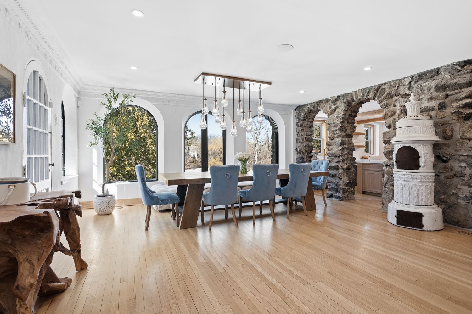 This elegant dining room features a large wooden table surrounded by plush, light blue upholstered chairs, illuminated by a striking multi-pendant chandelier. The space is defined by its unique architectural blend of smooth white walls with arched windows and a rustic, exposed stone wall with arched openings leading to the kitchen. A vintage-style white ceramic stove stands as a decorative focal point, creating a sophisticated yet warm atmosphere.