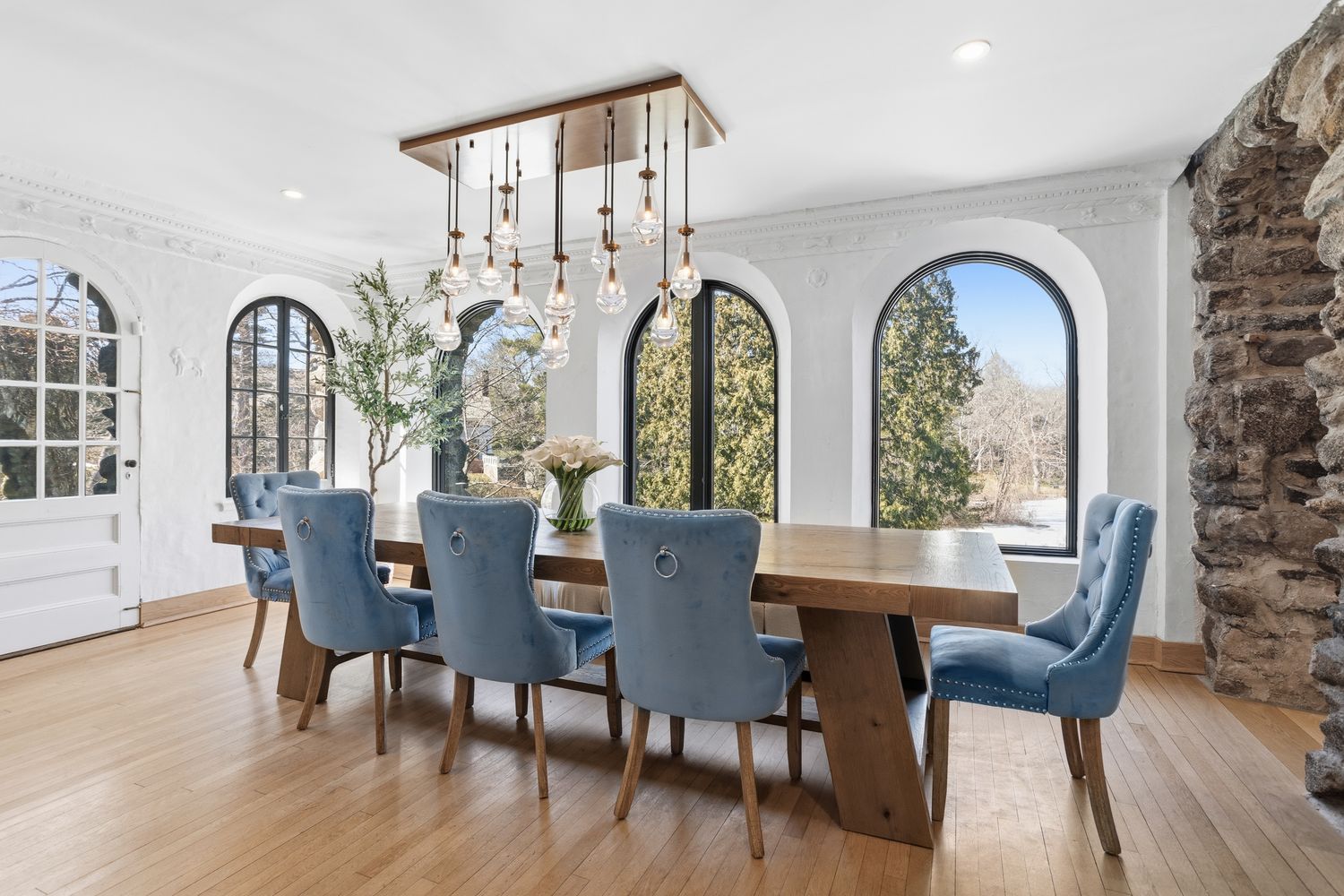 This elegant dining room features a large, rustic wooden table surrounded by six plush, light blue velvet chairs with nailhead trim and ring-pull details. The space is illuminated by a striking multi-pendant light fixture suspended from a wooden canopy, set against a backdrop of arched windows and a textured stone wall. The room exudes a sophisticated, warm atmosphere, blending traditional architectural elements with modern, luxurious furnishings.