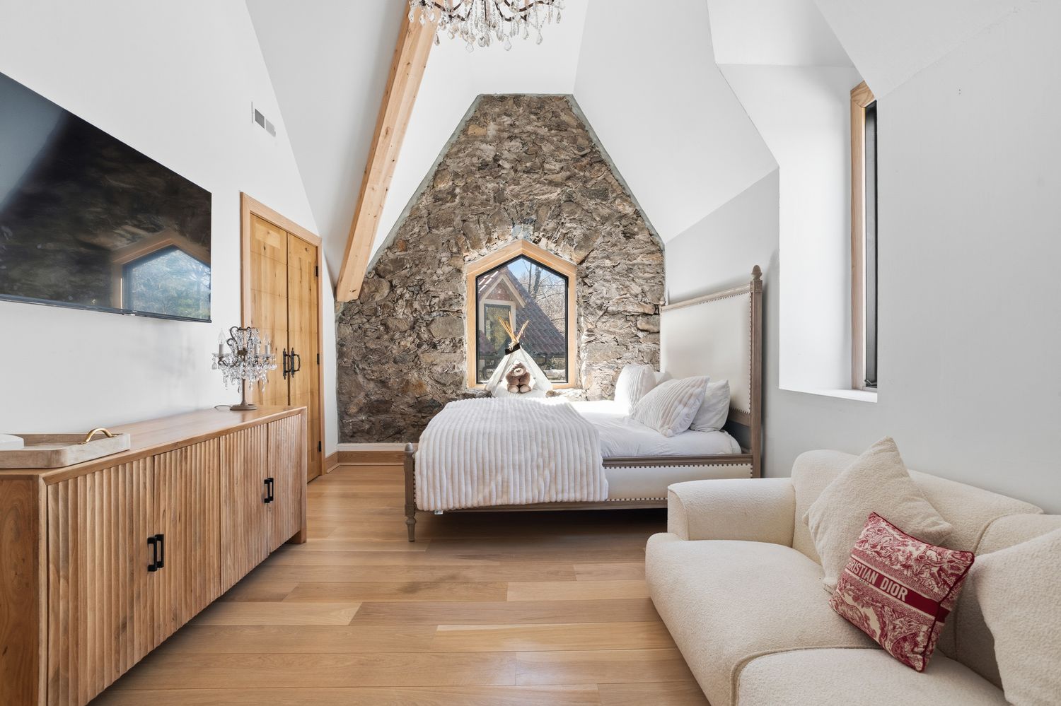This charming bedroom features a unique stone accent wall with a pointed arch window, creating a rustic yet elegant atmosphere. The room is furnished with a plush bed, a textured wooden dresser, and a cozy cream-colored sofa, all set against light wood flooring and vaulted ceilings. The space is bright and airy, blending modern comfort with architectural character.