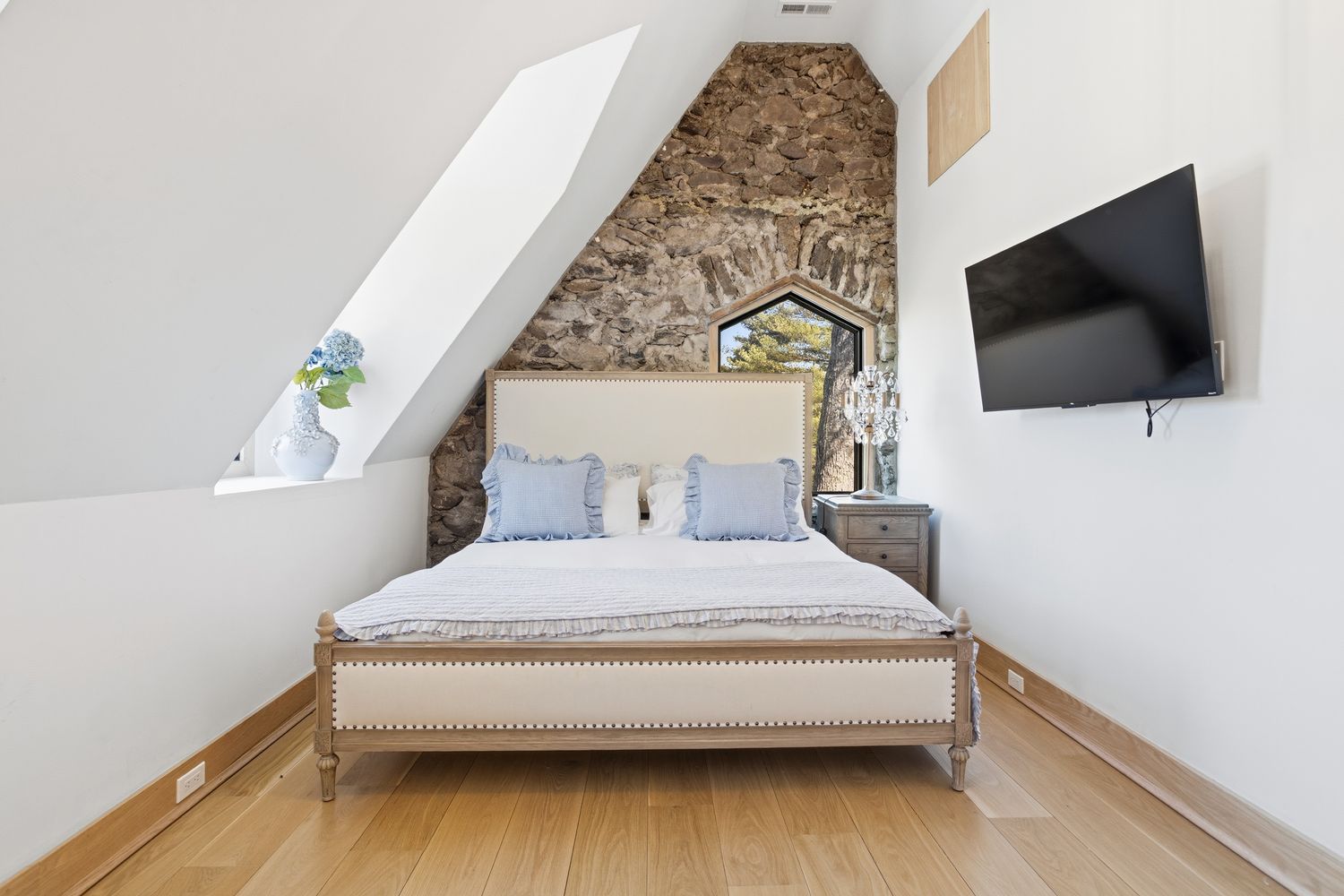 This charming bedroom features a unique architectural design with a sloped ceiling and a striking stone accent wall that frames a gothic-style window. The room is furnished with a light-colored upholstered bed, a matching nightstand with a crystal lamp, and a wall-mounted television, creating a cozy and rustic-chic atmosphere. The light hardwood flooring and bright white walls enhance the airy feel of this attic-style space.