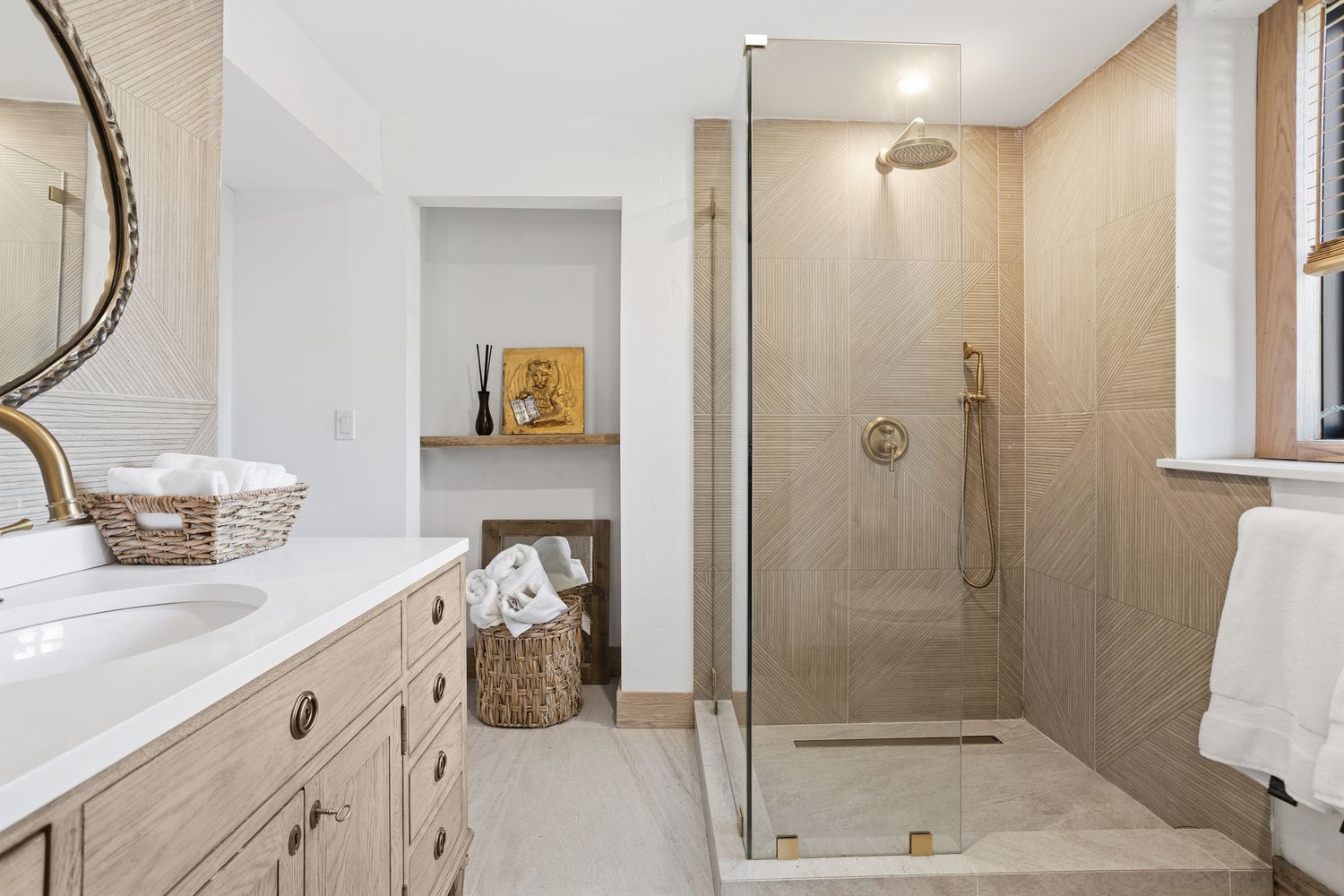 This elegant primary bathroom features a light wood vanity with a white countertop and a walk-in shower with textured, geometric-patterned wall tiles. The space is accented with warm brass fixtures, a round mirror, and natural woven baskets, creating a serene and spa-like atmosphere. The perspective is a straight-on shot capturing the seamless transition between the vanity area and the glass-enclosed shower.