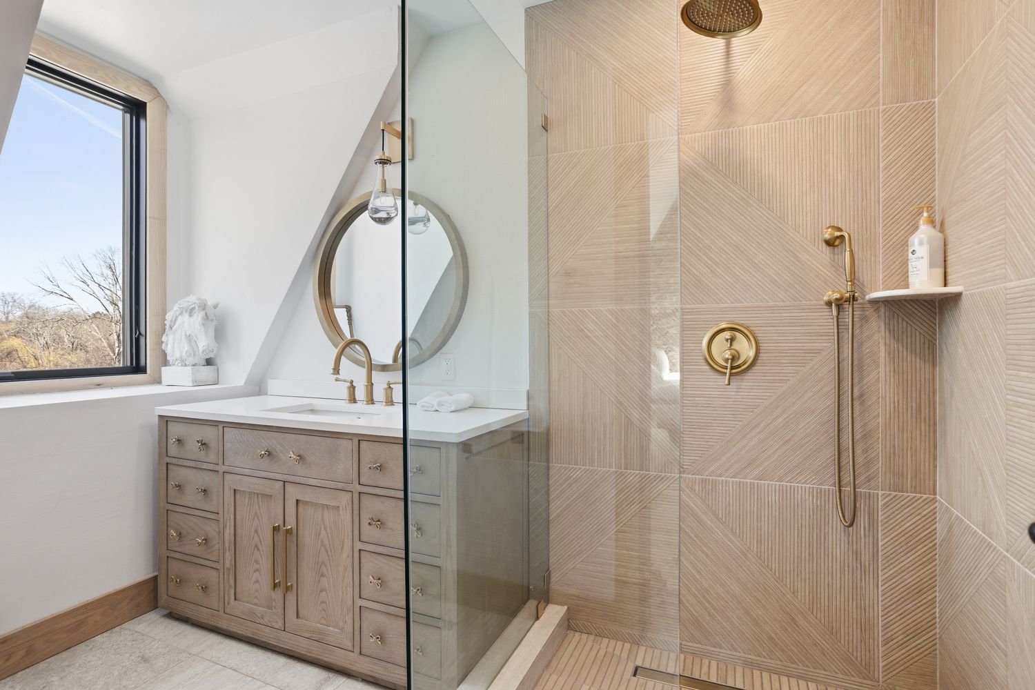 This elegant primary bathroom features a custom wood vanity with a white quartz countertop and a chic circular brass-framed mirror. A spacious walk-in shower is enclosed by a frameless glass panel, showcasing sophisticated textured wall tiles and high-end brass fixtures, including a rainfall showerhead. The room is bathed in natural light from a large window, creating a bright, luxurious, and spa-like atmosphere.