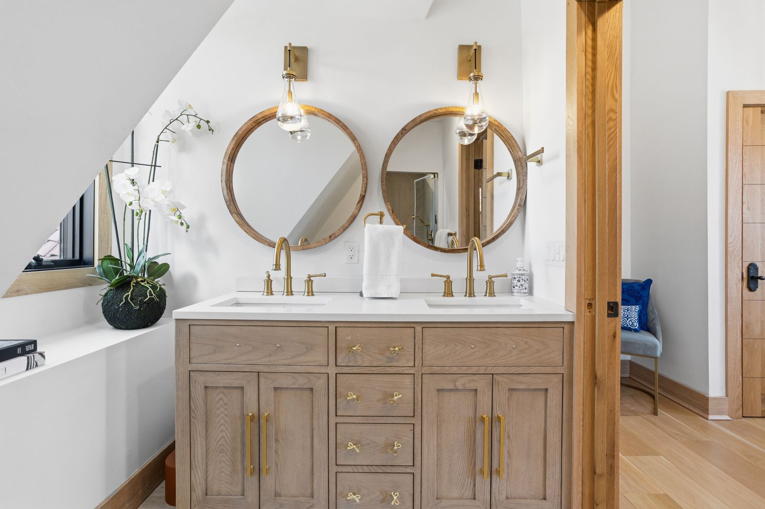 This elegant primary bathroom features a double vanity with light oak cabinetry, gold hardware, and a clean white countertop. Two circular mirrors are mounted above the sinks, complemented by stylish wall-mounted light fixtures and a potted orchid on the side ledge. The space exudes a warm, modern aesthetic with high-quality finishes and a bright, inviting atmosphere.