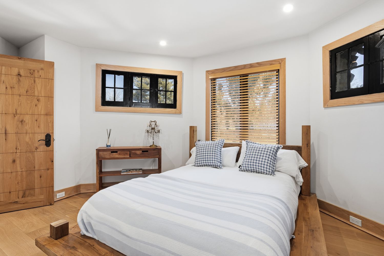 This cozy bedroom features a rustic aesthetic with a wooden platform bed frame, matching wood trim, and a natural wood door. The room is brightened by white walls and multiple windows with black frames, while a small wooden console table adds a touch of charm. The overall impression is one of warm, inviting comfort with a blend of modern and farmhouse design elements.