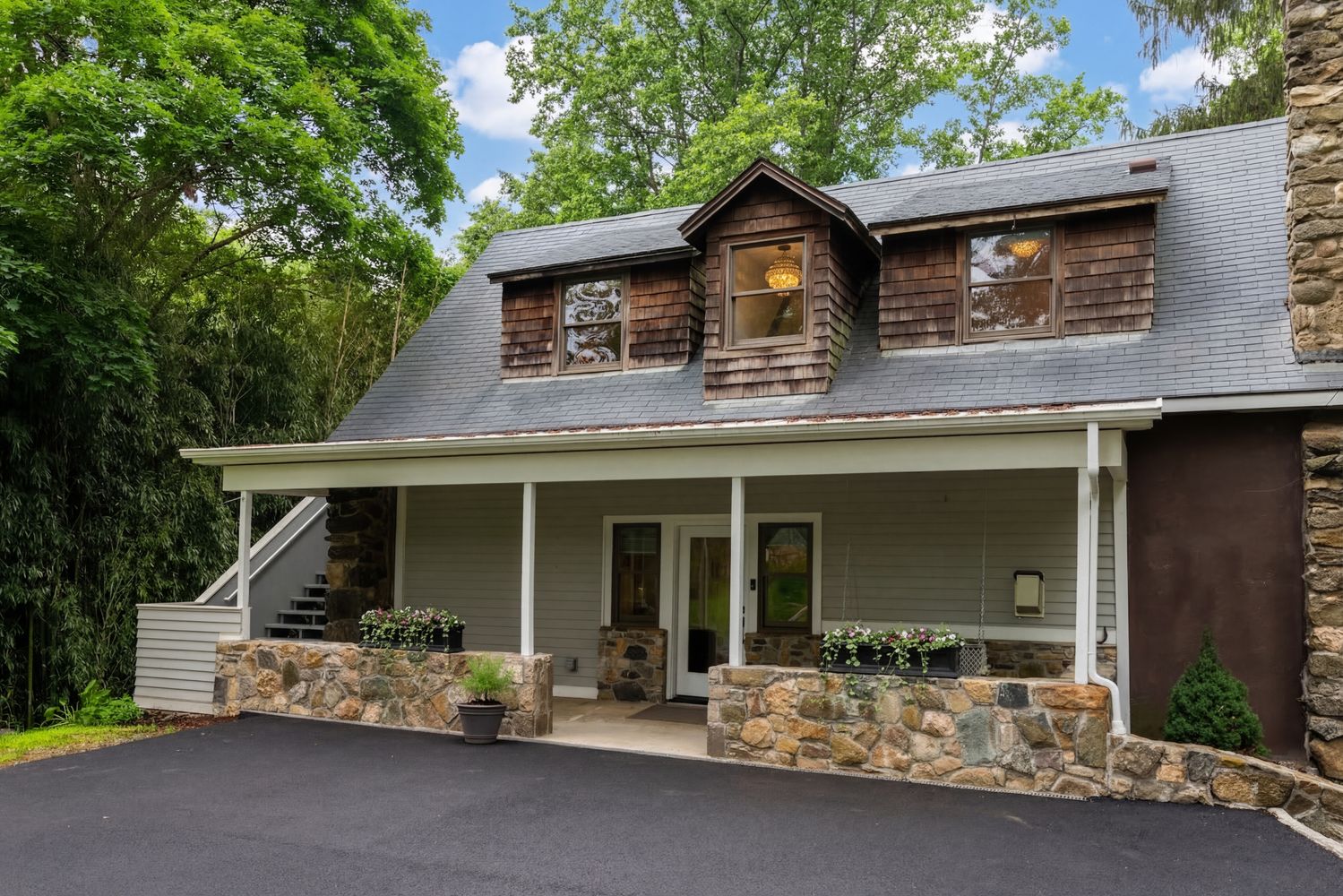 This charming cottage-style home features a classic blend of stone masonry and wood shingle siding, accented by a welcoming covered porch. The exterior is framed by lush greenery and a paved driveway, with dormer windows adding character to the roofline. The overall aesthetic is rustic and inviting, suggesting a cozy, well-maintained residence.