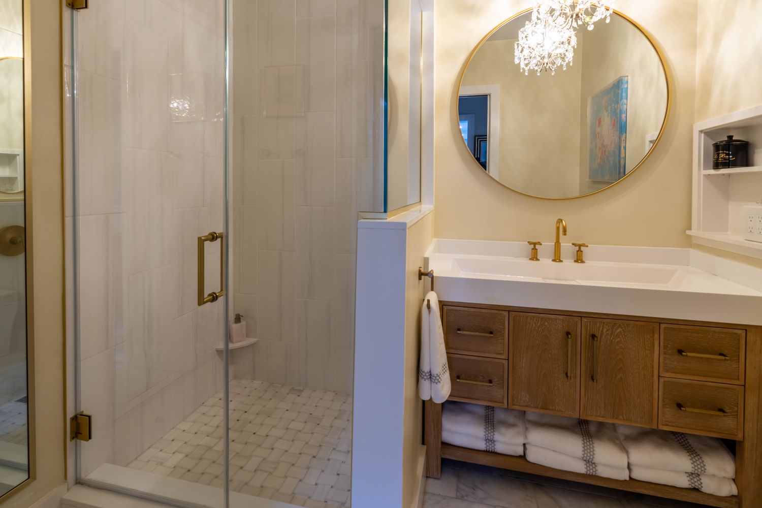This elegant primary bathroom features a walk-in shower with white subway tile and a glass enclosure, adjacent to a custom light oak vanity with a white countertop and gold fixtures. A large, round gold-framed mirror hangs above the vanity, reflecting a crystal chandelier, while plush white towels are neatly stored on the open lower shelf. The space exudes a sophisticated, spa-like atmosphere with a warm and inviting color palette.