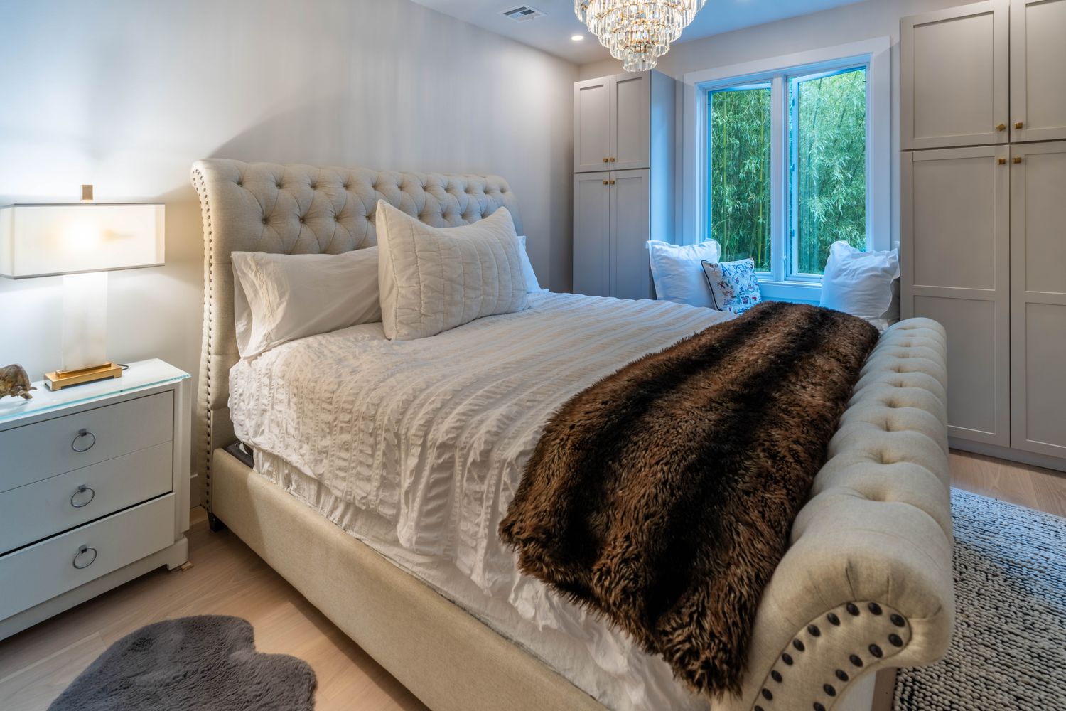This elegant primary bedroom features a tufted, upholstered sleigh bed as the centerpiece, adorned with a plush faux-fur throw and textured white bedding. The room is framed by built-in cabinetry on either side of a window seat, creating a cozy nook that overlooks lush greenery outside. A crystal chandelier adds a touch of luxury, while the neutral color palette and soft lighting contribute to a serene and sophisticated atmosphere.
