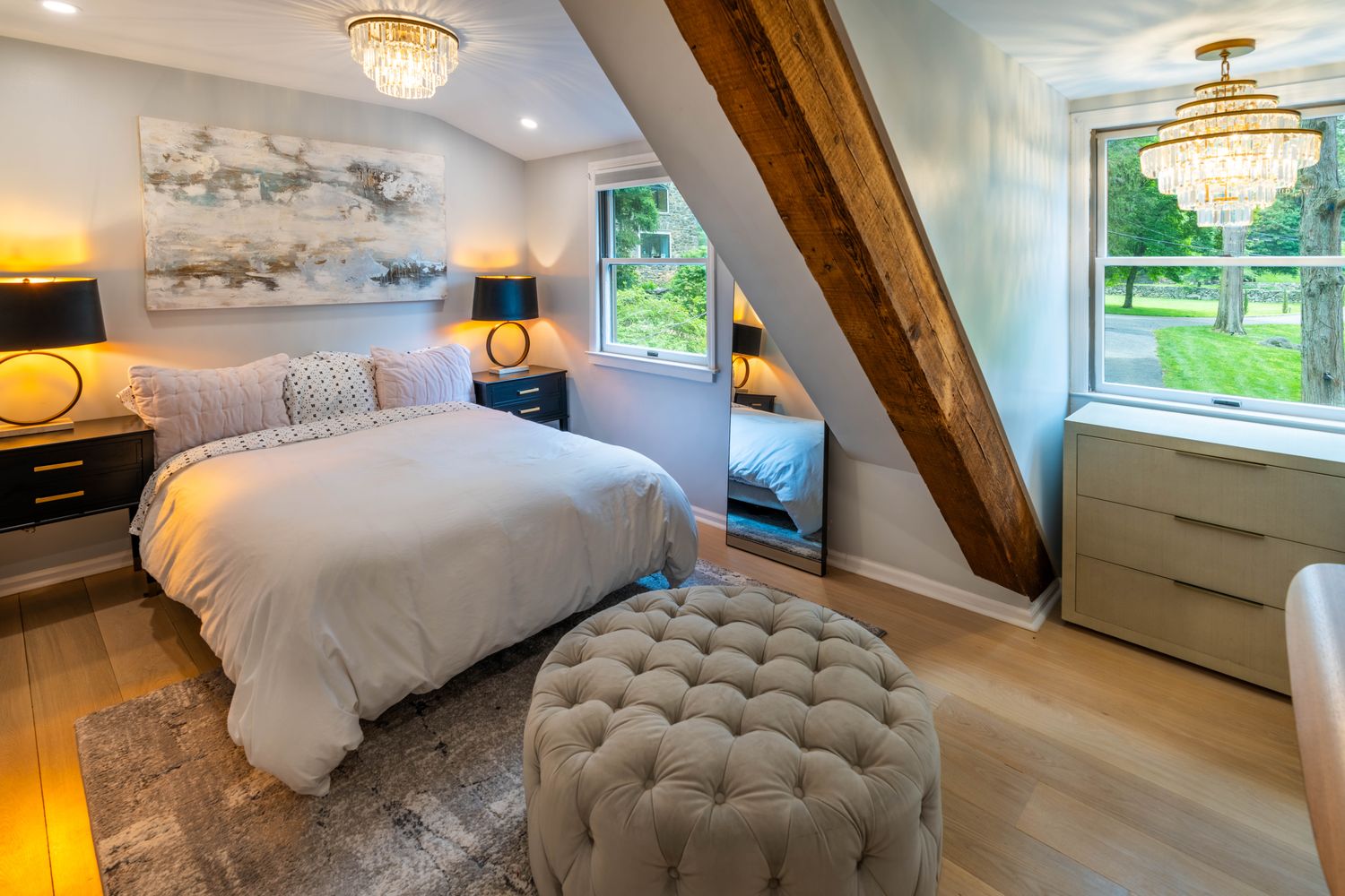 This elegant primary bedroom features a cozy, tufted ottoman at the foot of a plush bed, set against a backdrop of soft gray walls and a striking exposed wooden ceiling beam. The room is illuminated by two sophisticated crystal chandeliers and warm bedside lamps, creating a serene and luxurious atmosphere. A large window provides a peaceful view of the lush green outdoors, while a full-length mirror adds depth to the space.