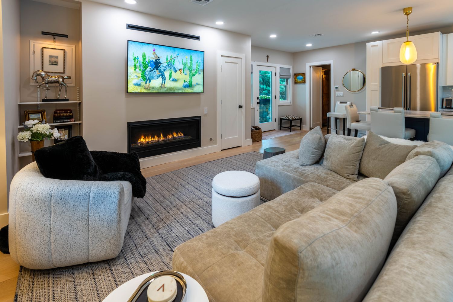 This inviting open-concept living room features a modern linear fireplace set beneath a wall-mounted television, creating a cozy focal point. The space is furnished with a plush, neutral-toned sectional sofa and a textured accent chair, all grounded by a large area rug that flows seamlessly into the adjacent kitchen area. The bright, airy atmosphere is enhanced by recessed lighting and a neutral color palette, making it an ideal space for relaxation and entertaining.