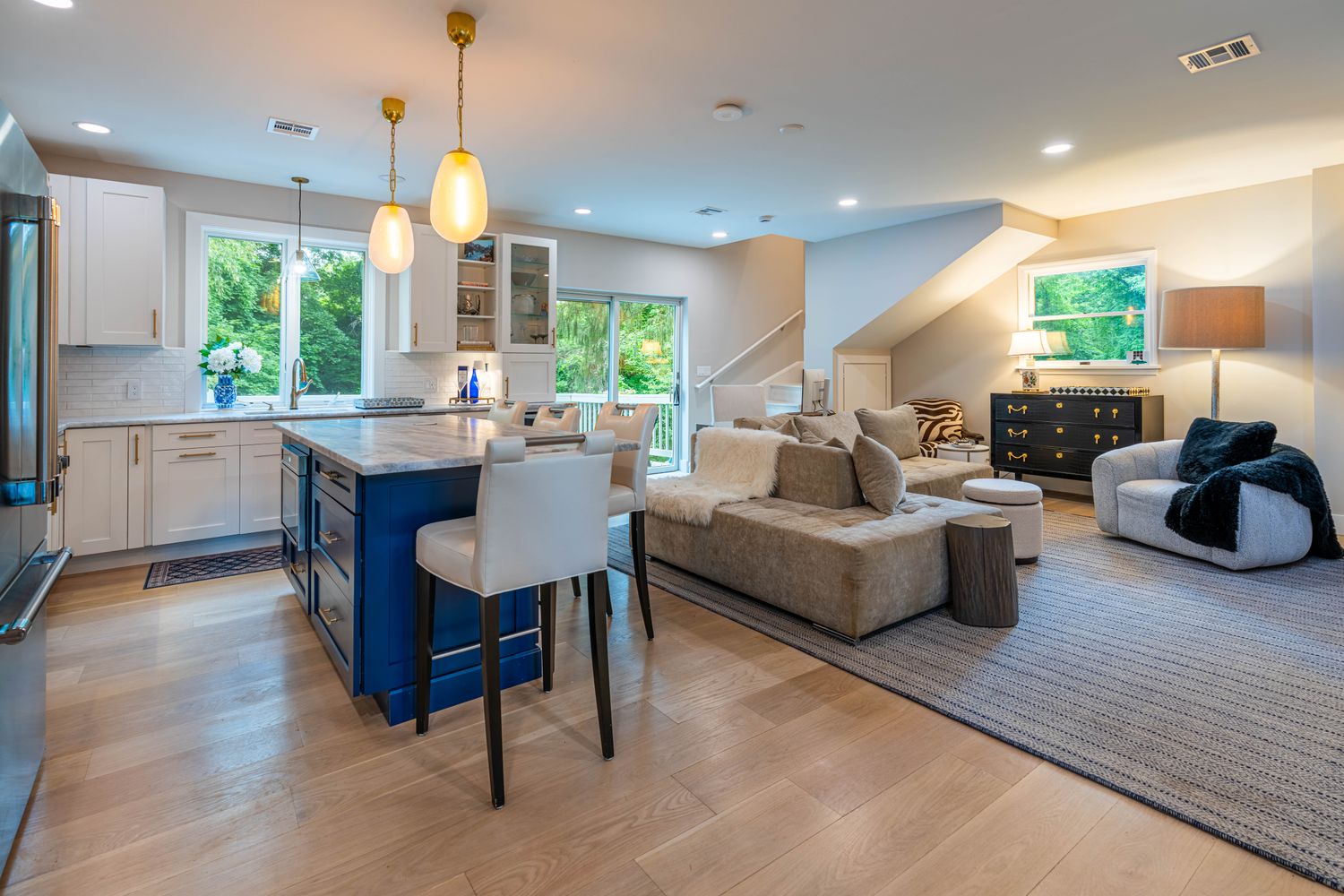 This open-concept living space seamlessly blends a modern kitchen with a cozy living area, featuring a striking blue kitchen island topped with marble-style countertops. The kitchen is equipped with white cabinetry and gold hardware, while the adjacent living room offers a comfortable sectional sofa, a textured area rug, and a stylish armchair. The space is bright and inviting, with large windows and glass sliding doors that provide ample natural light and a view of the outdoors.