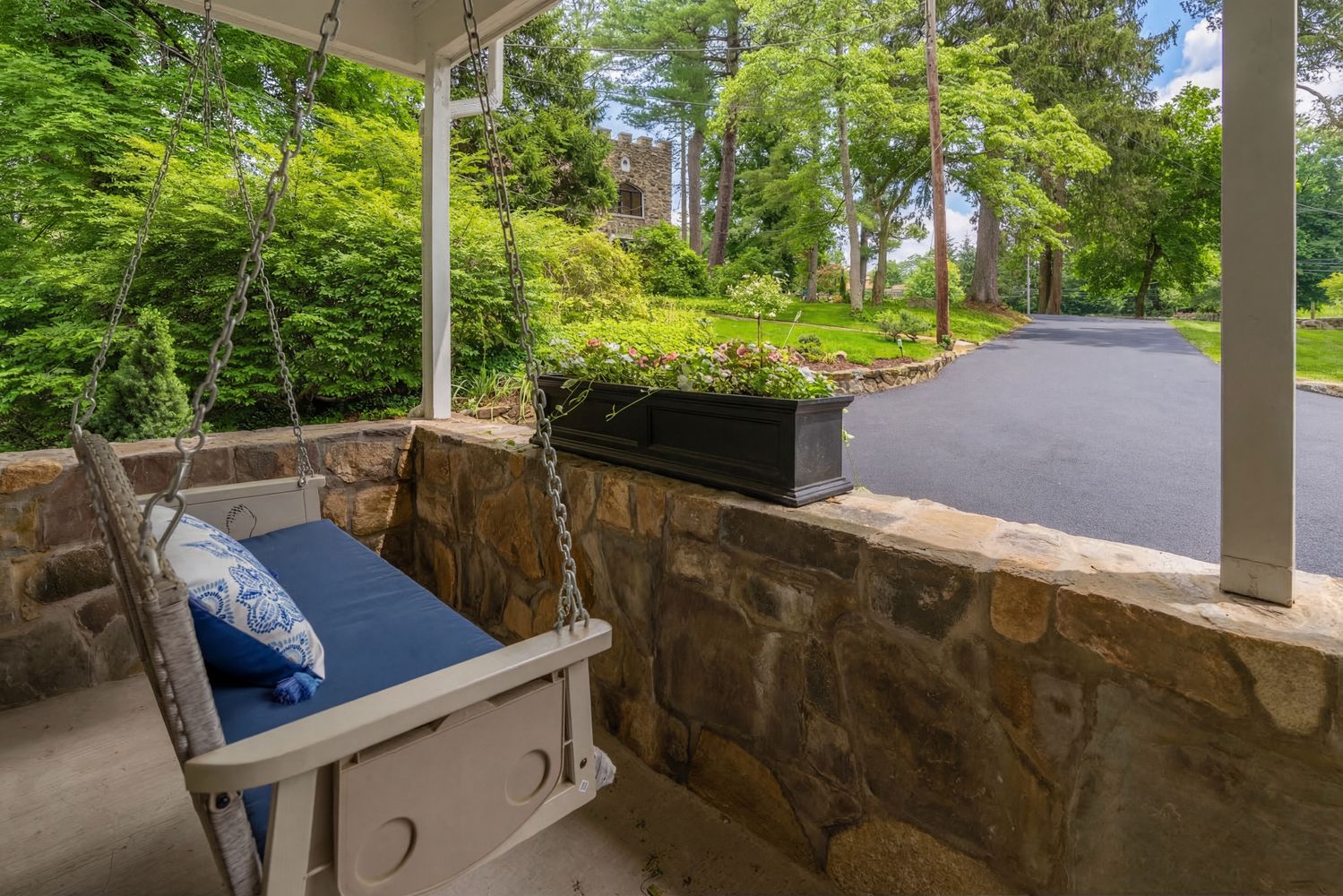 This inviting front porch features a classic hanging swing with a plush blue cushion, set against a rustic stone wall. A black planter box filled with vibrant flowers adds a touch of color, while the view overlooks a paved driveway leading toward a charming stone structure nestled among lush trees. The perspective captures a serene, peaceful atmosphere perfect for relaxing outdoors.