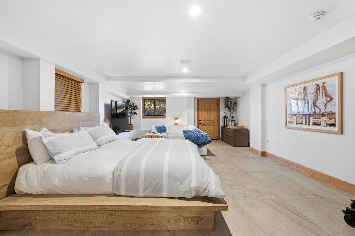 This spacious primary bedroom features a large platform bed with a natural wood headboard and light blue striped bedding, creating a serene and modern aesthetic. The room seamlessly transitions into a cozy seating area with a white sofa and a television, while the neutral color palette and light-toned flooring enhance the airy, open feel. A large framed photograph on the wall adds a touch of personality to this well-lit, contemporary living space.